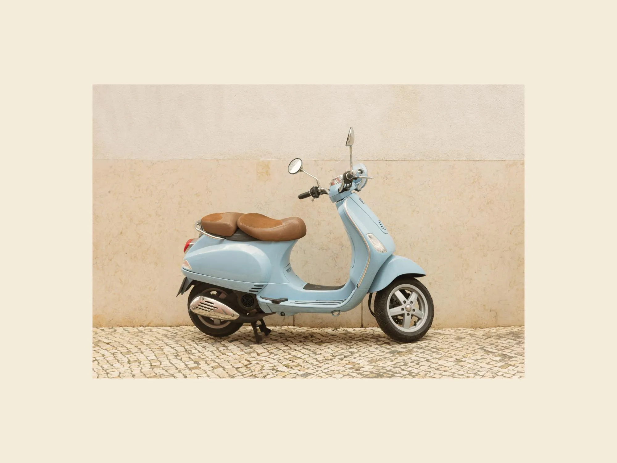 Poster einer hellblauen Vespa mit brauner Sitzbank, geparkt auf Kopfsteinpflaster vor einer beigen Wand.