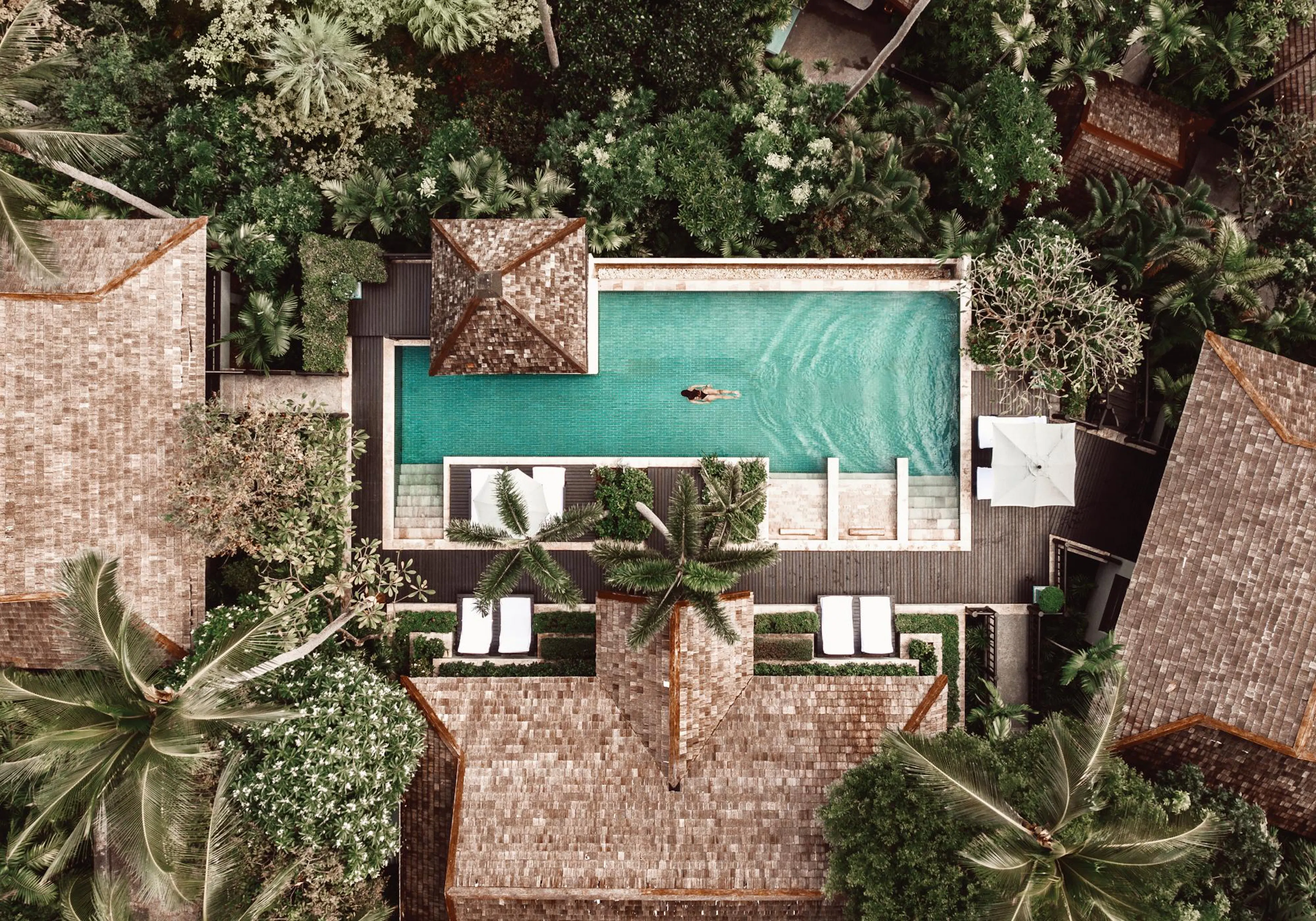 Poster: Eine Luftaufnahme einer Hotelanlage mit blauem Pool und schwimmender Person, umgeben von grünen Bäumen und Dächern.
