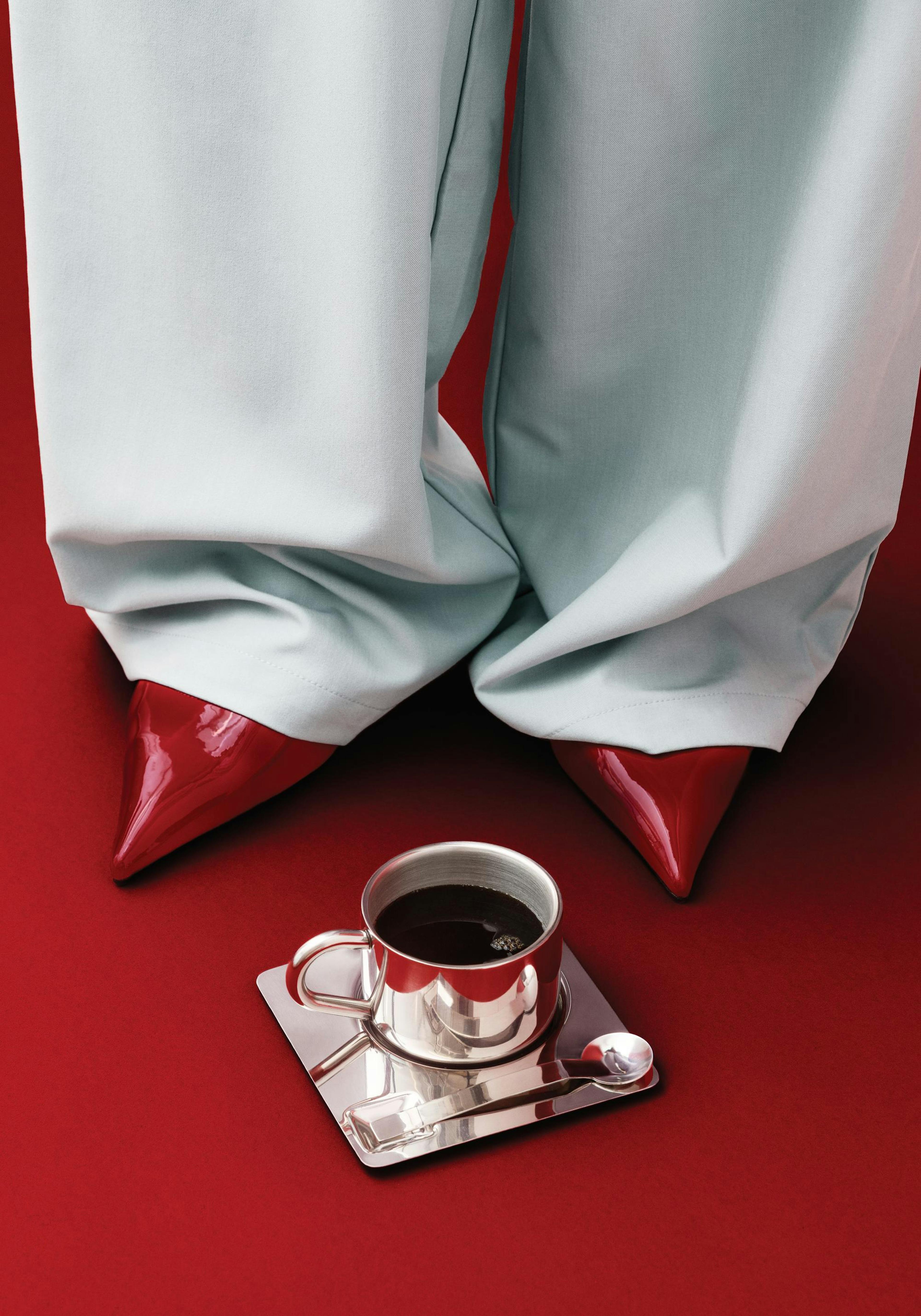 Affiche dune personne en pantalon gris clair et chaussures à talons rouges, avec une tasse de café argentée au premier plan sur 