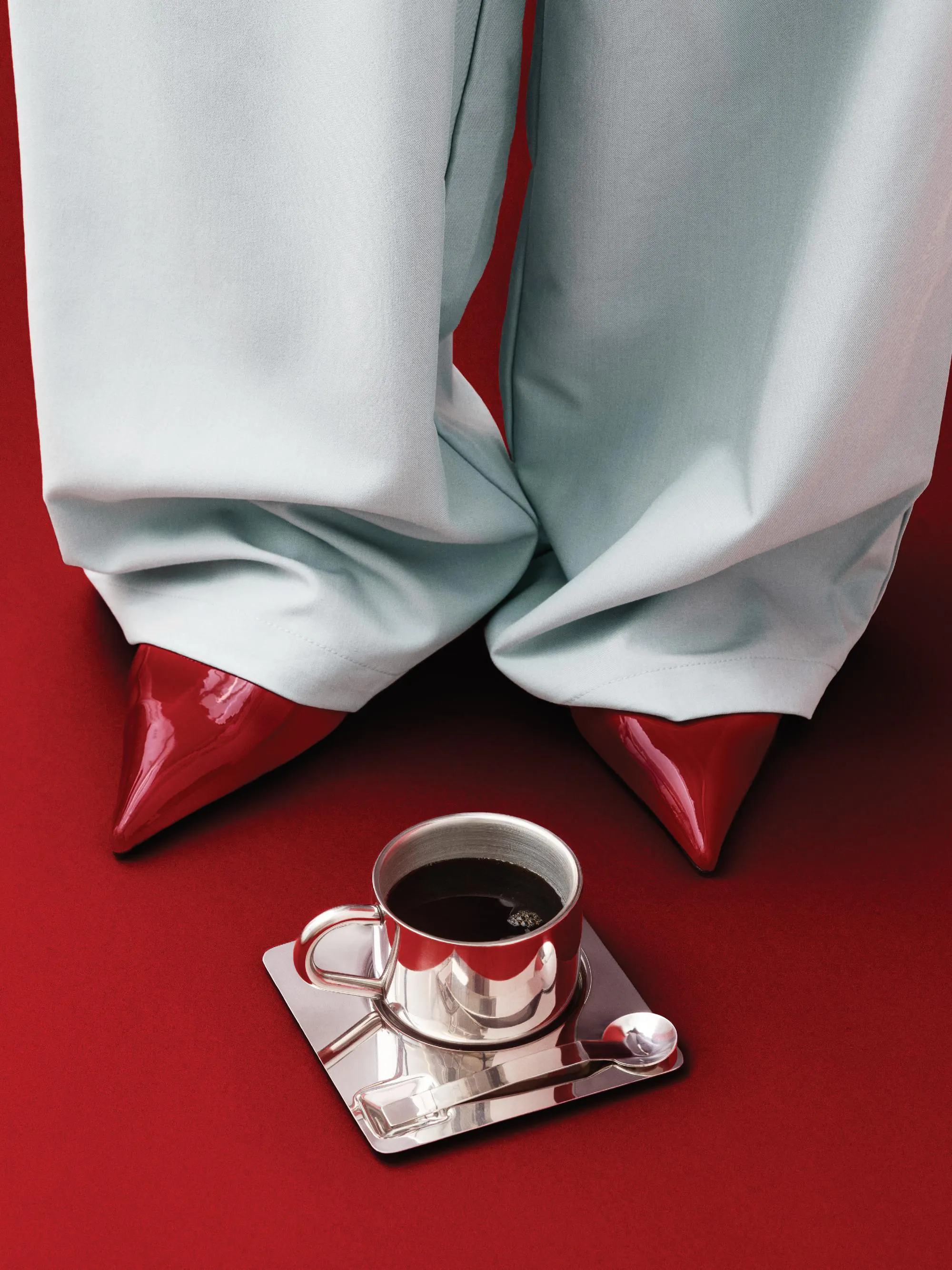 Ένα poster με γυναικεία πόδια με γαλάζιο παντελόνι και κόκκινες γόβες, δίπλα σε ένα φλιτζάνι καφέ σε κόκκινο φόντο.