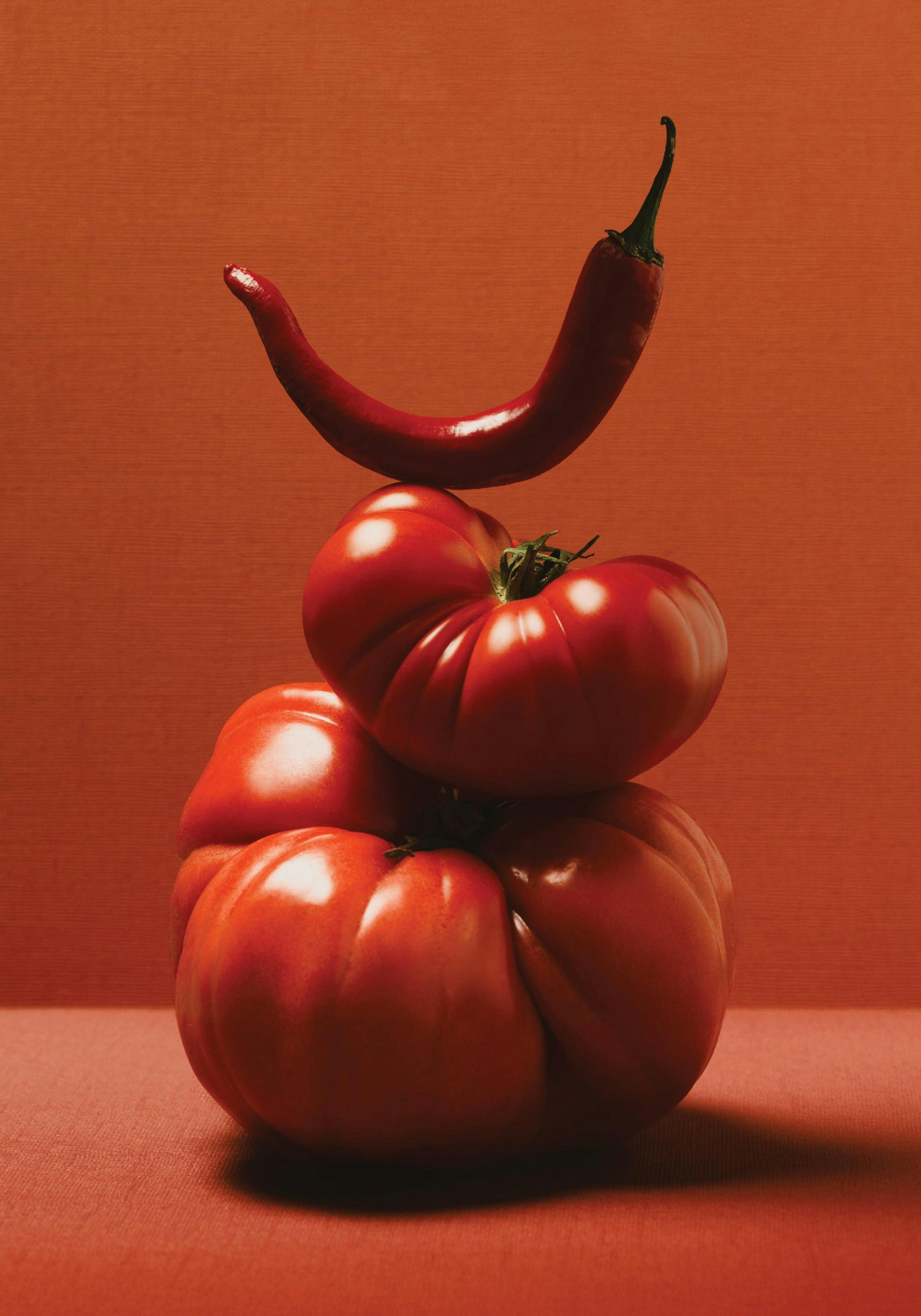 A still life print of a red chili pepper balancing on two stacked tomatoes on a red background.