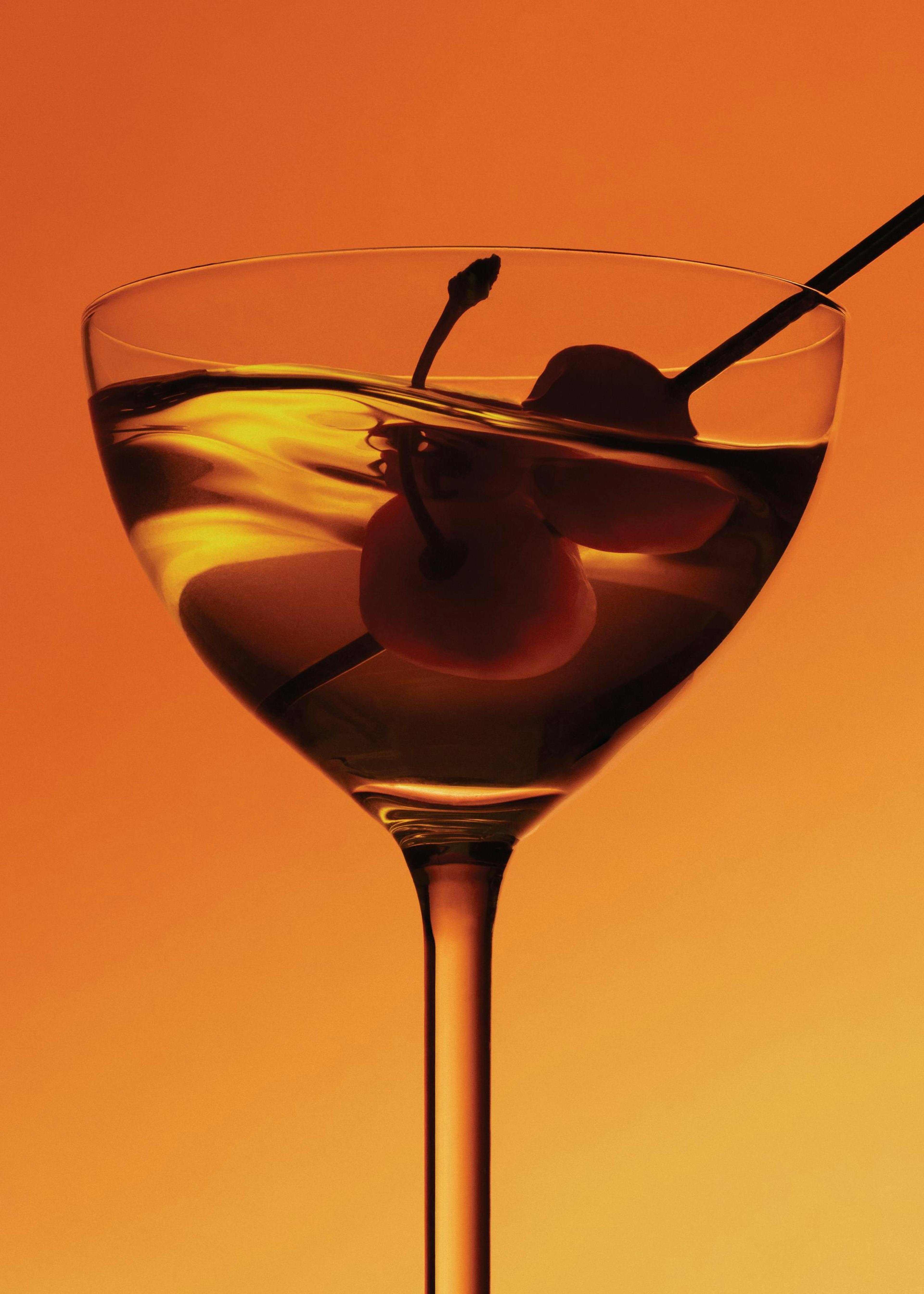 Póster de un cóctel dorado con dos cerezas y una pajita en copa de martini, sobre fondo naranja.