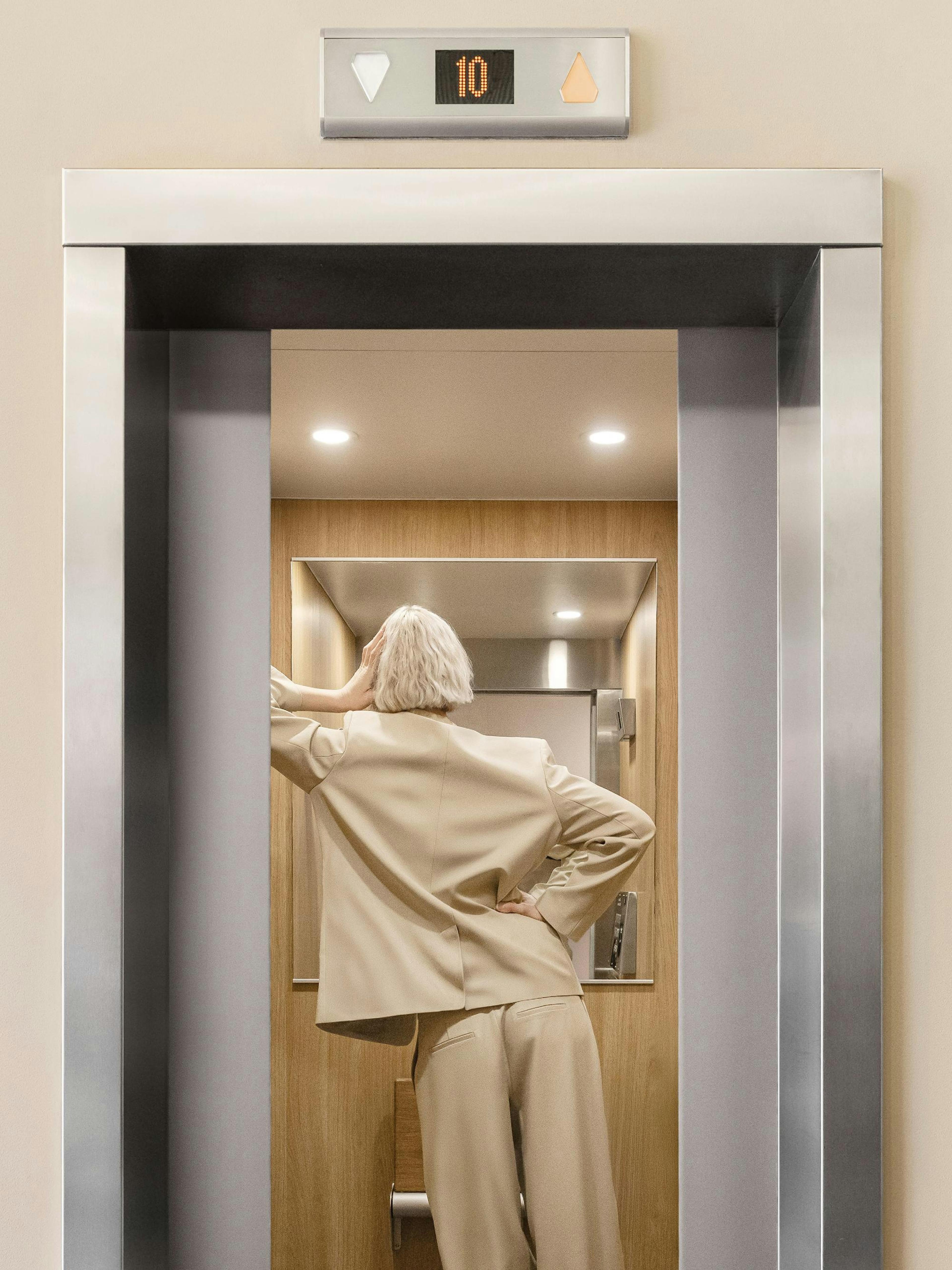 A print of a woman with blonde hair in a beige suit stepping into an open elevator with a digital display showing 10.