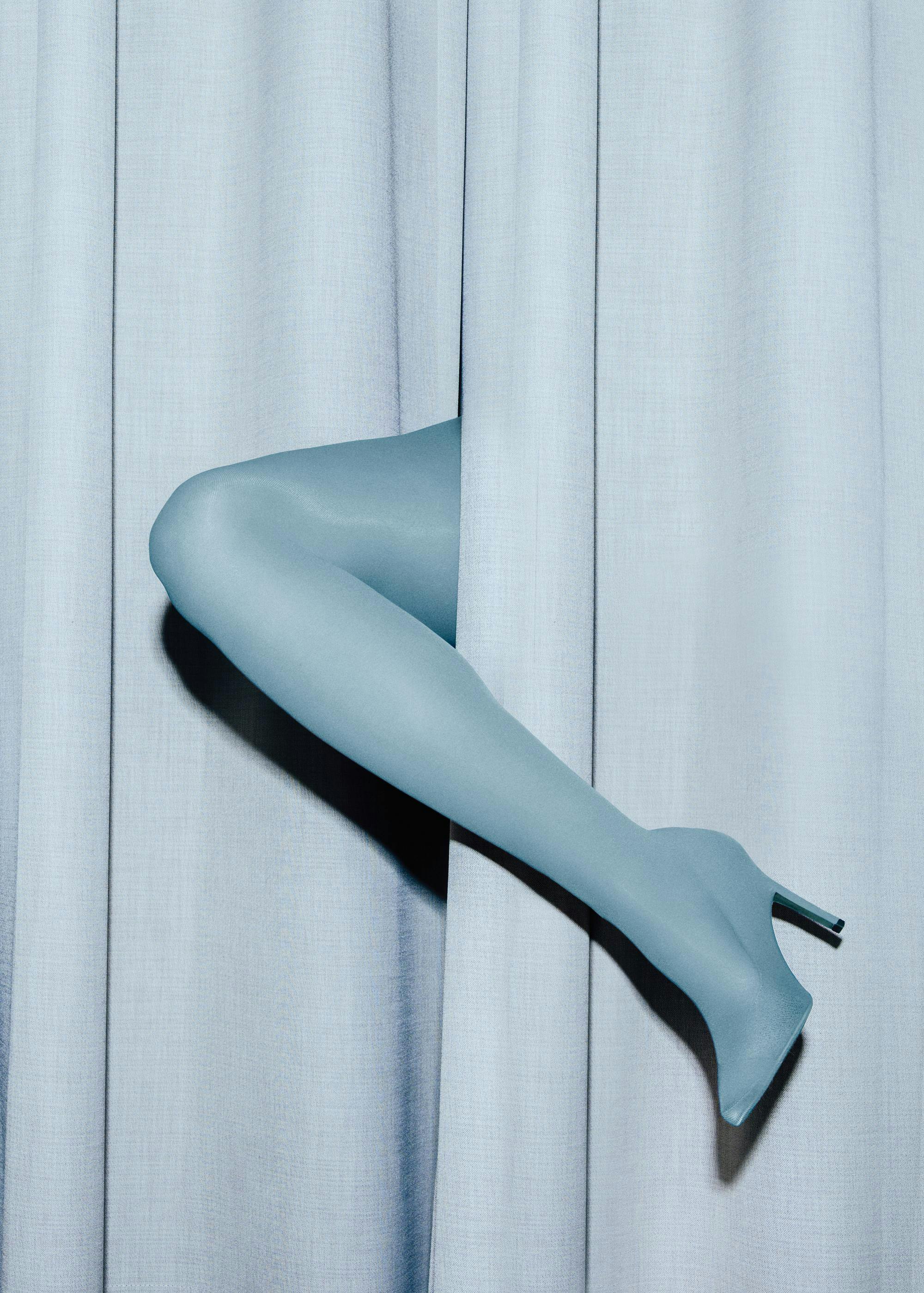 A print of a mannequins leg in a blue stocking and high heel, peeking from behind a light blue curtain.