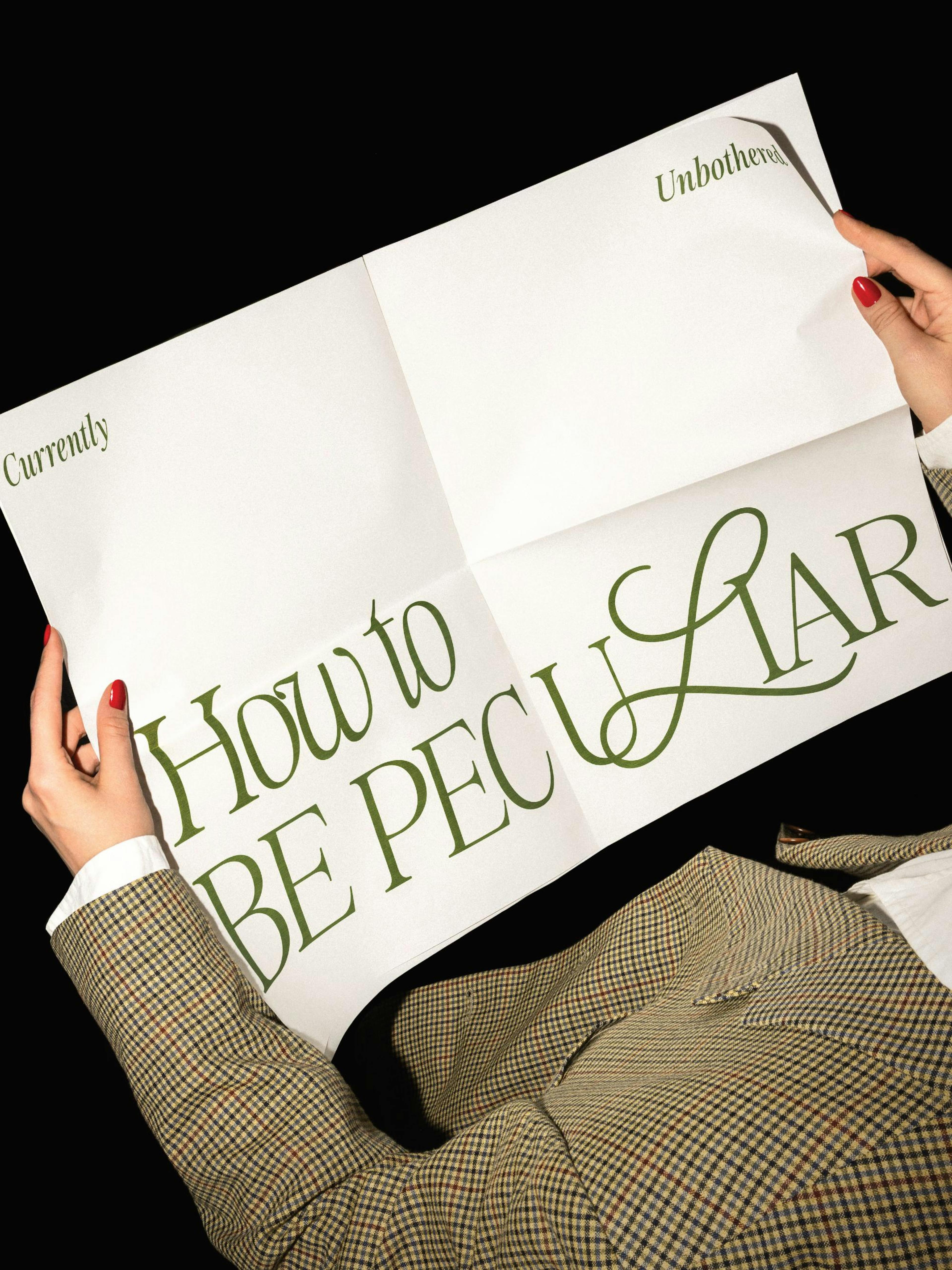 A print with the green text How to BE PECULIAR across the center, held by hands with red nails and a checkered jacket.