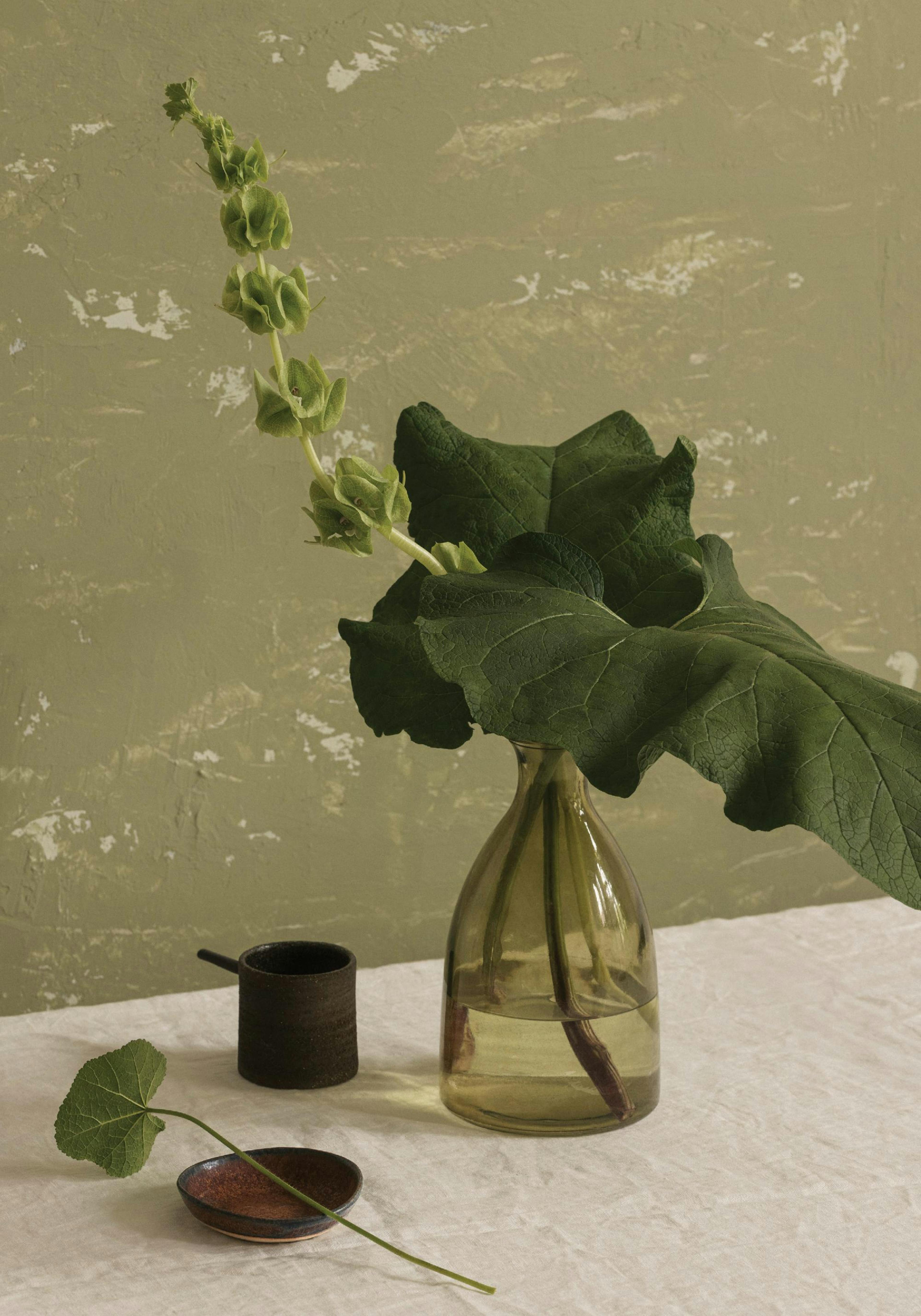 Un póster de un jarrón de vidrio con una rama verde de flores y hojas grandes sobre una mesa blanca.