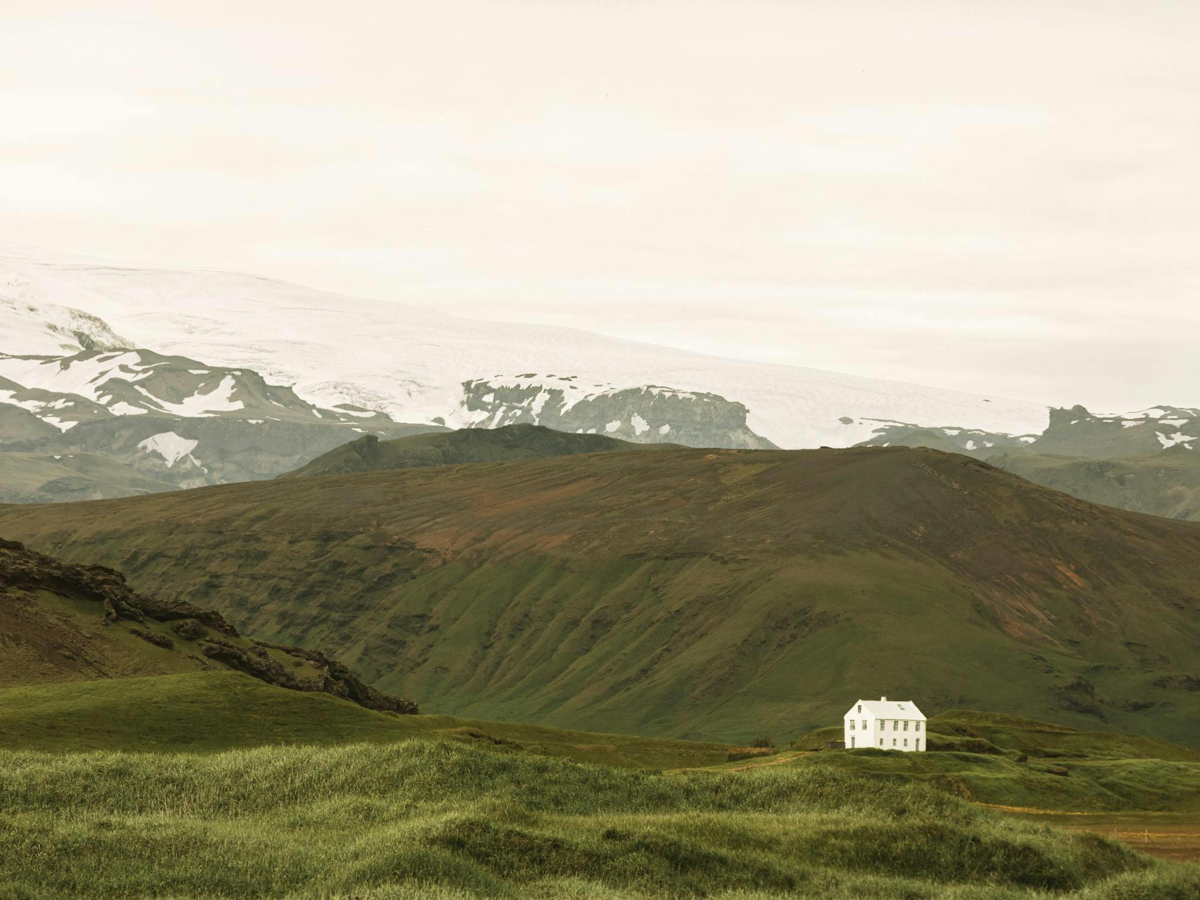 A print of a white house on a green hill with snow-capped mountains and a gray sky.