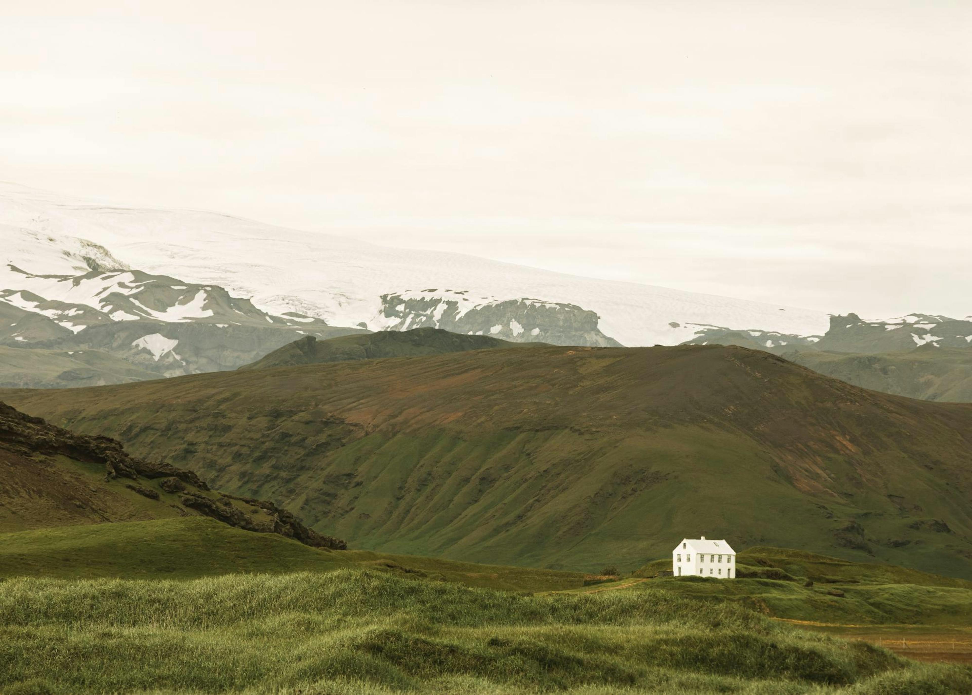 A print of a lone white house on a green hill with a snow-capped mountain range in the background under a cloudy sky.