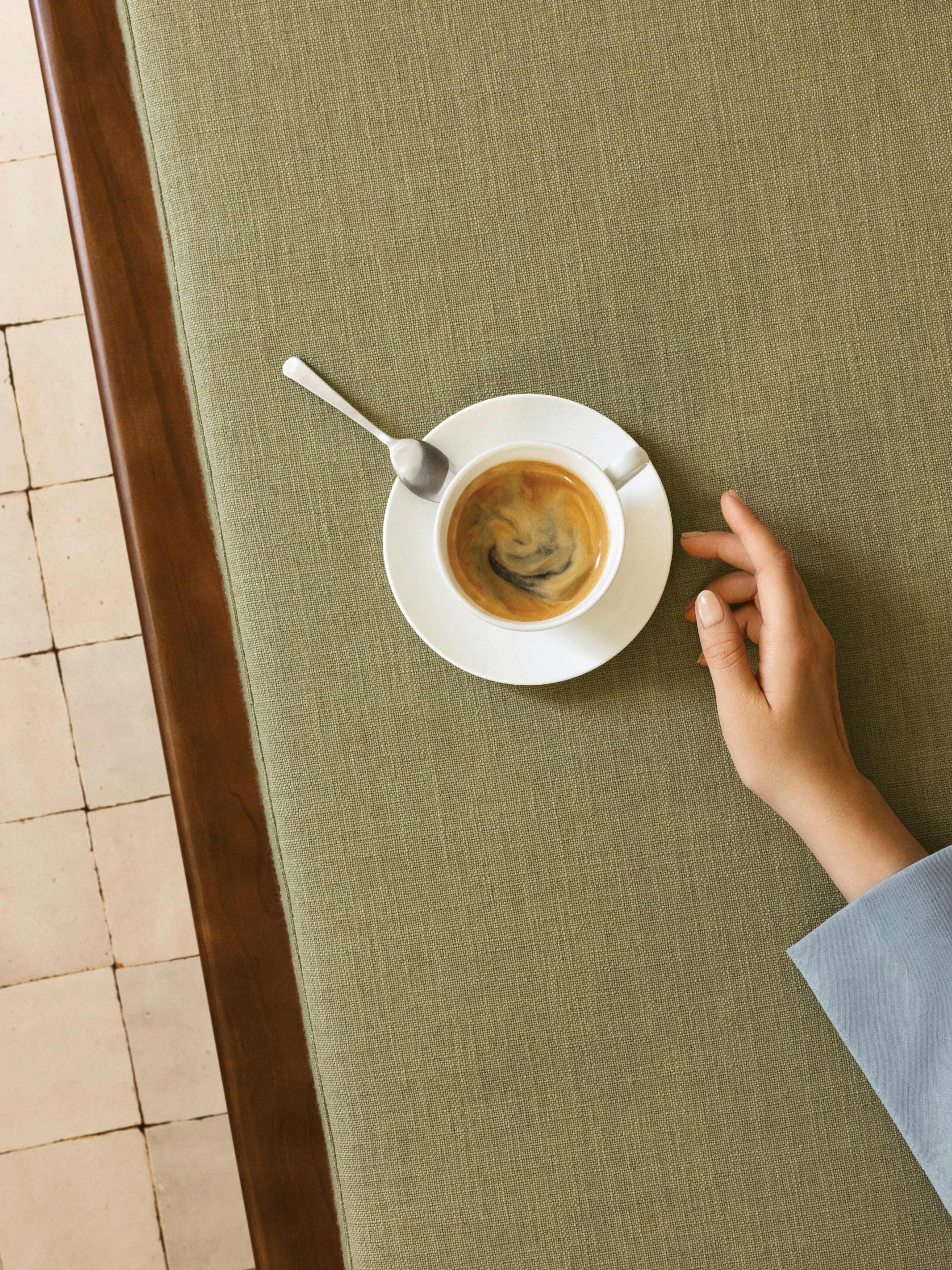Un póster de una taza de café espresso en una mesa verde con una mano tocándola.