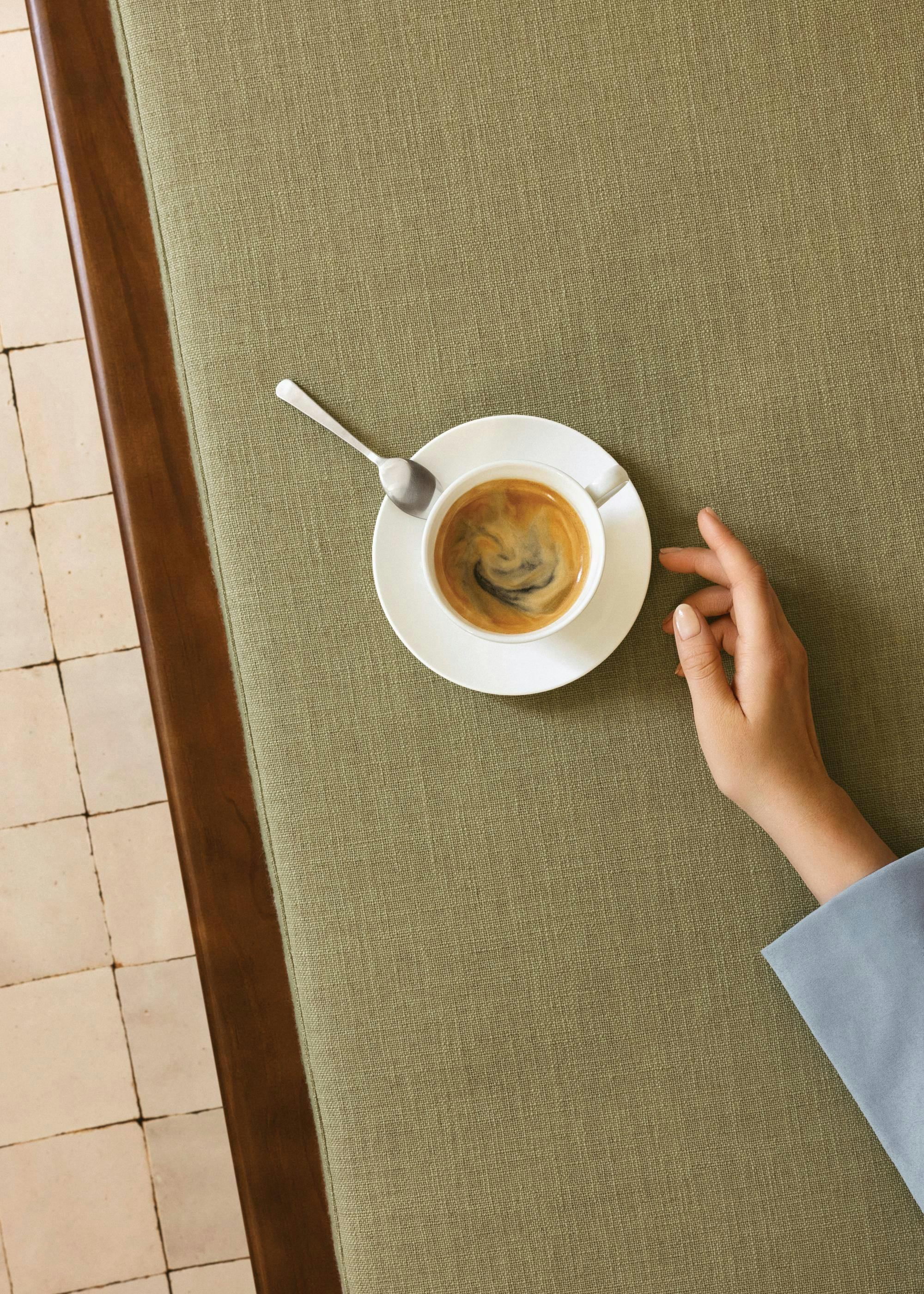 A print of an overhead view of a hand resting next to a white coffee cup and spoon on a textured green surface.