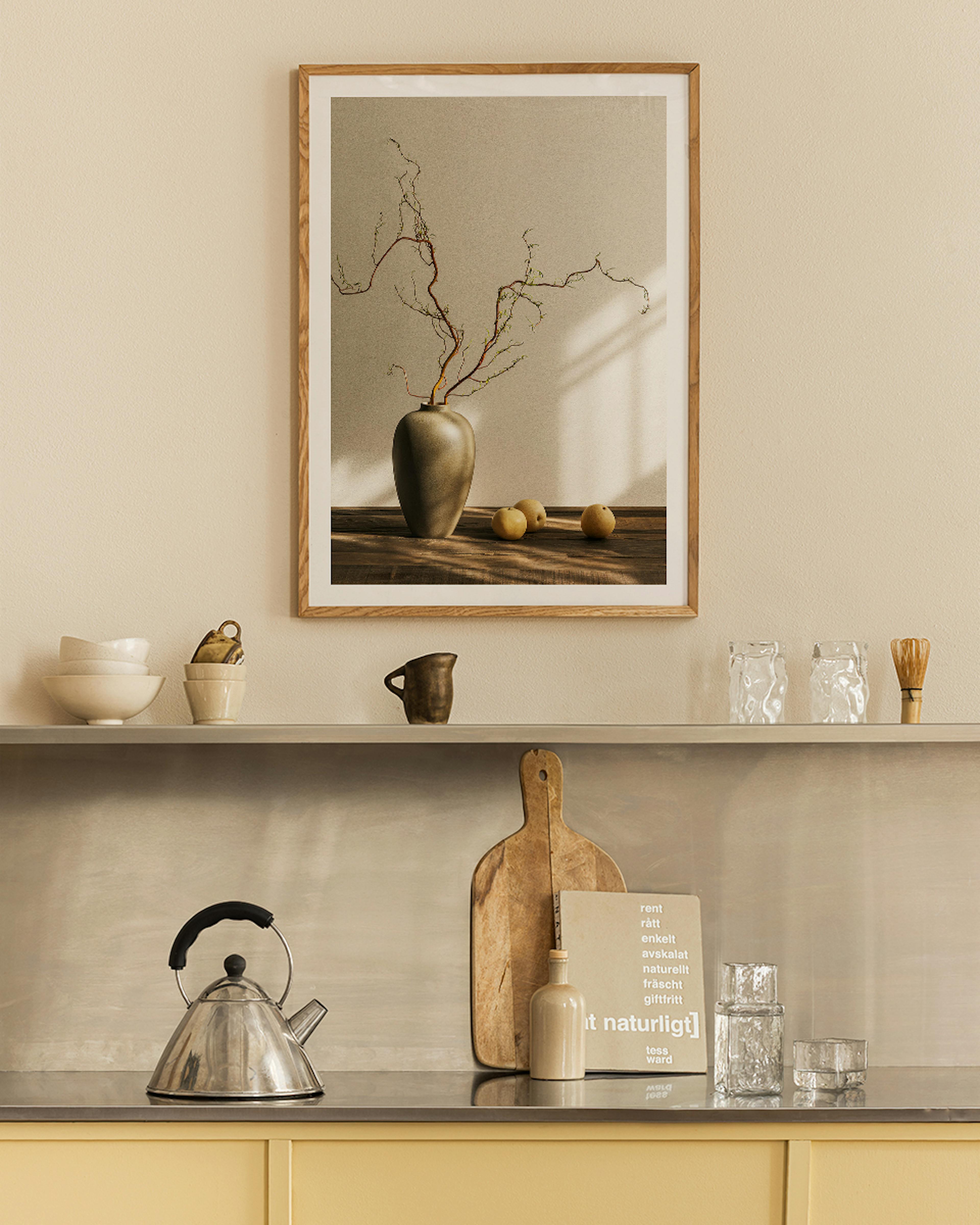 A print of a dark vase with branches and three light-colored fruits on a wooden surface, hanging above a kitchen shelf.