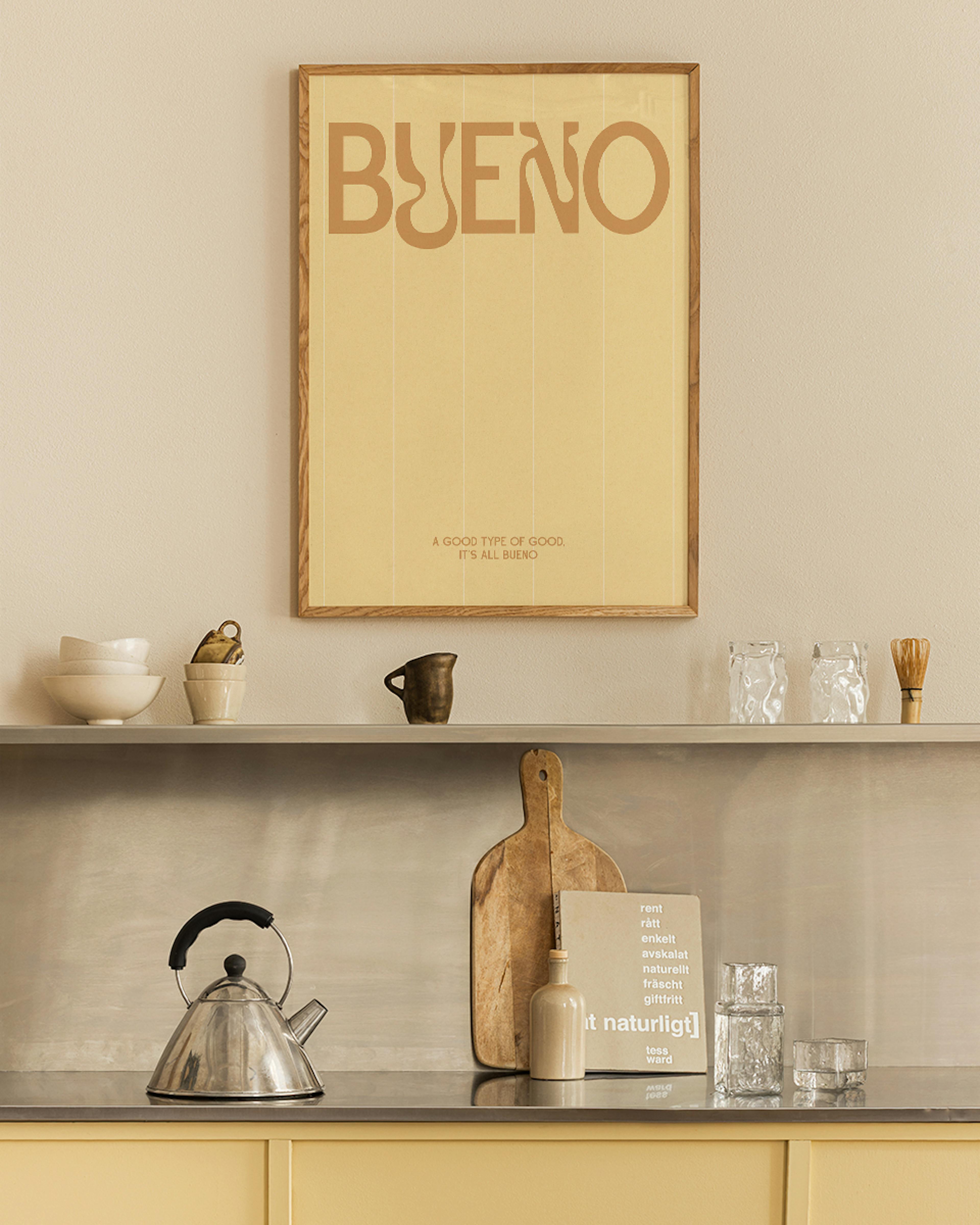 Póster amarillo crema con la palabra BUENO en marrón, colgado sobre un estante de cocina con utensilios.