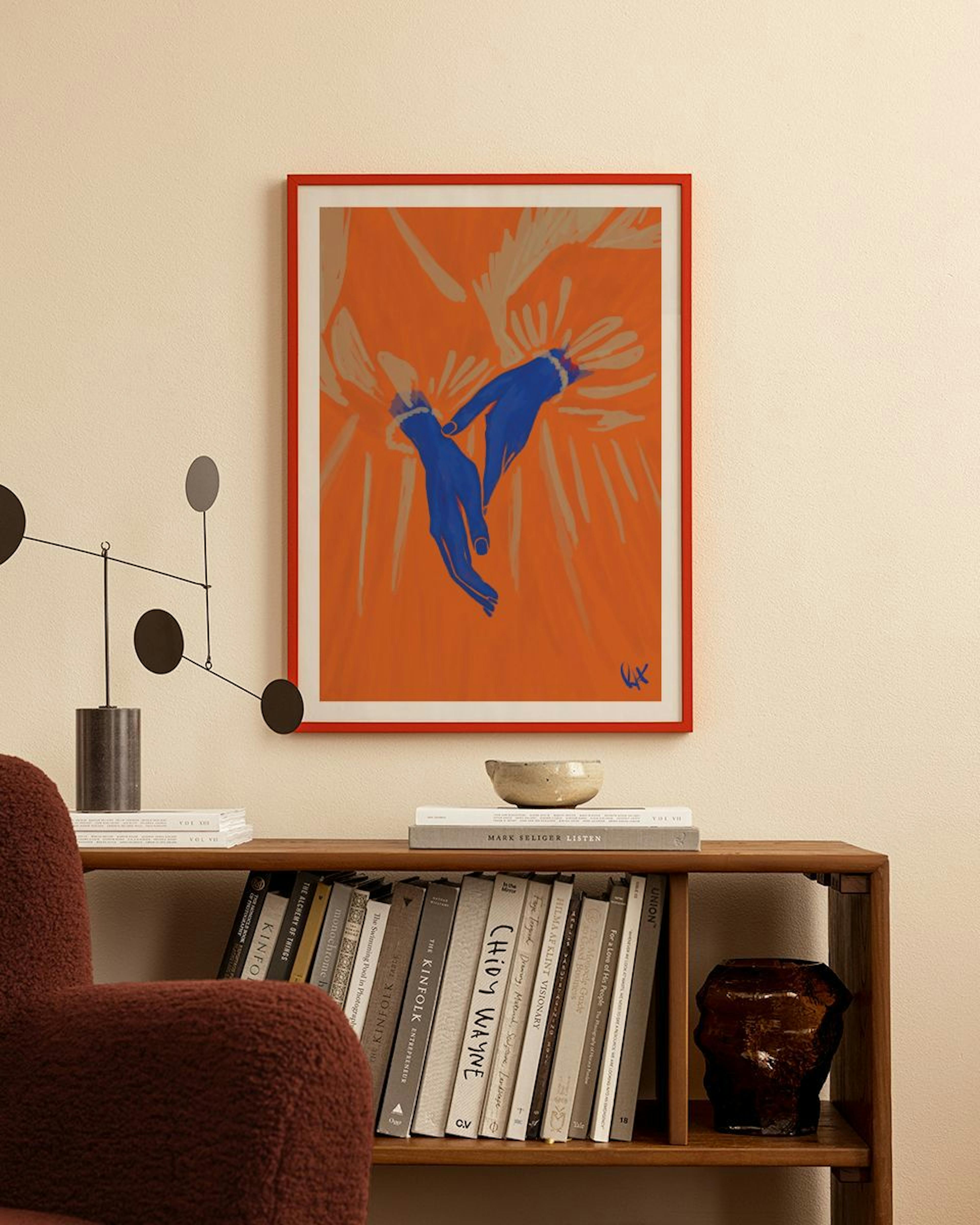 A print of blue hands reaching out on an orange and beige background, hanging above a wooden bookshelf in a cozy room.
