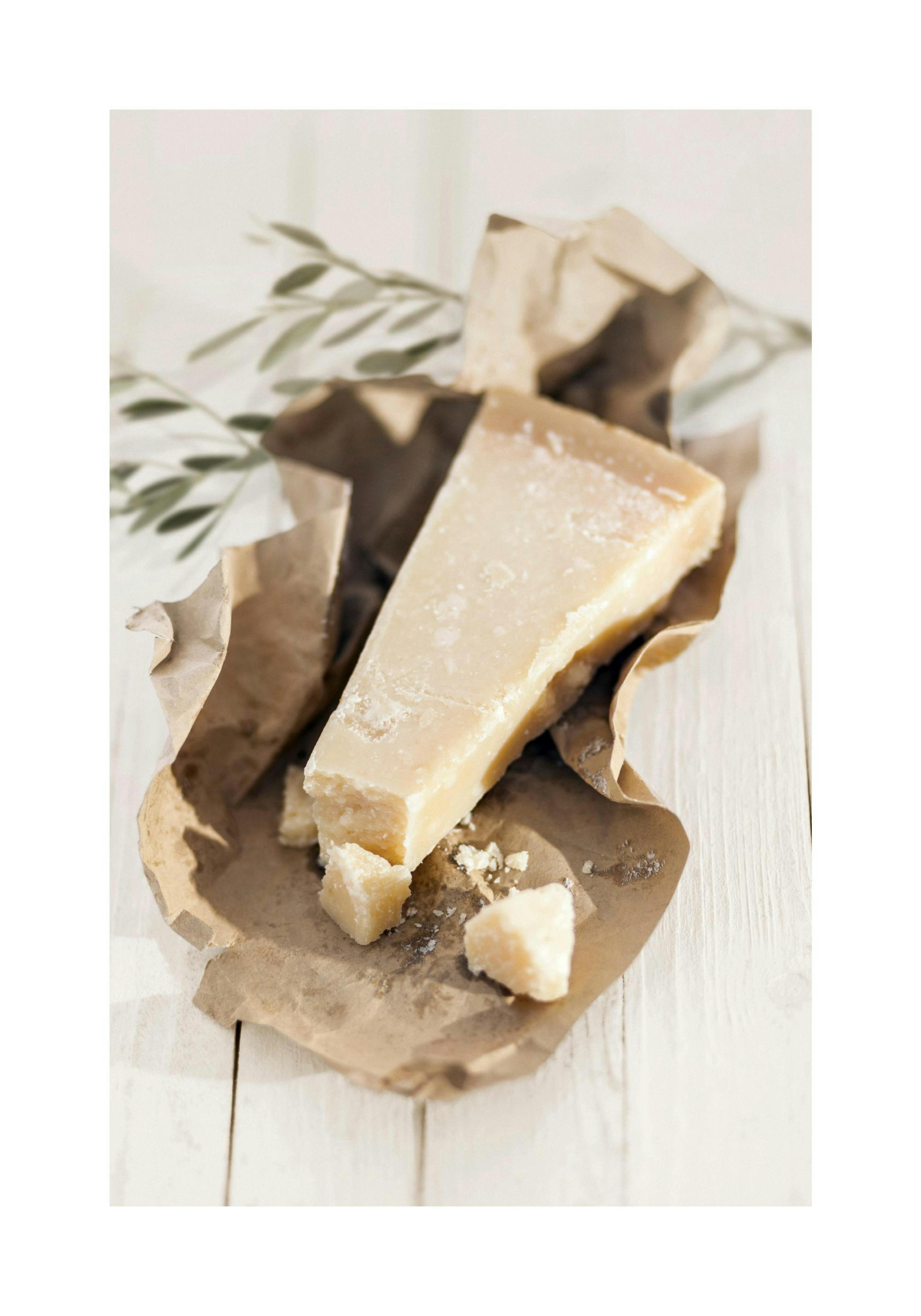 Beige valokuva parmesaanijuustosta ruskeassa paperissa valkoisella pöydällä. Oliivinoksa taustalla.