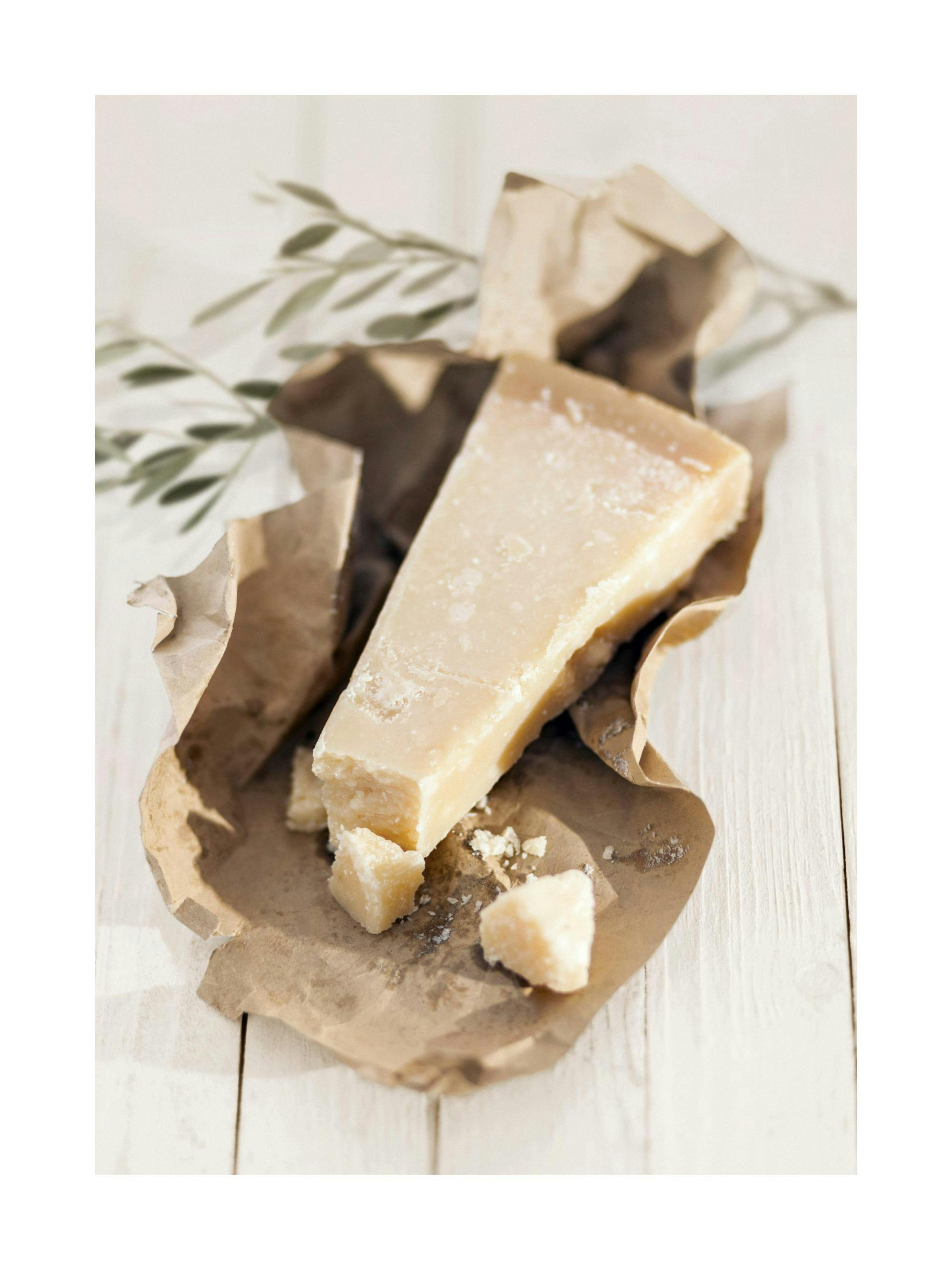 Fotografi av en bit parmesanost på brunt papper med olivkvist på ett vitt träbord.