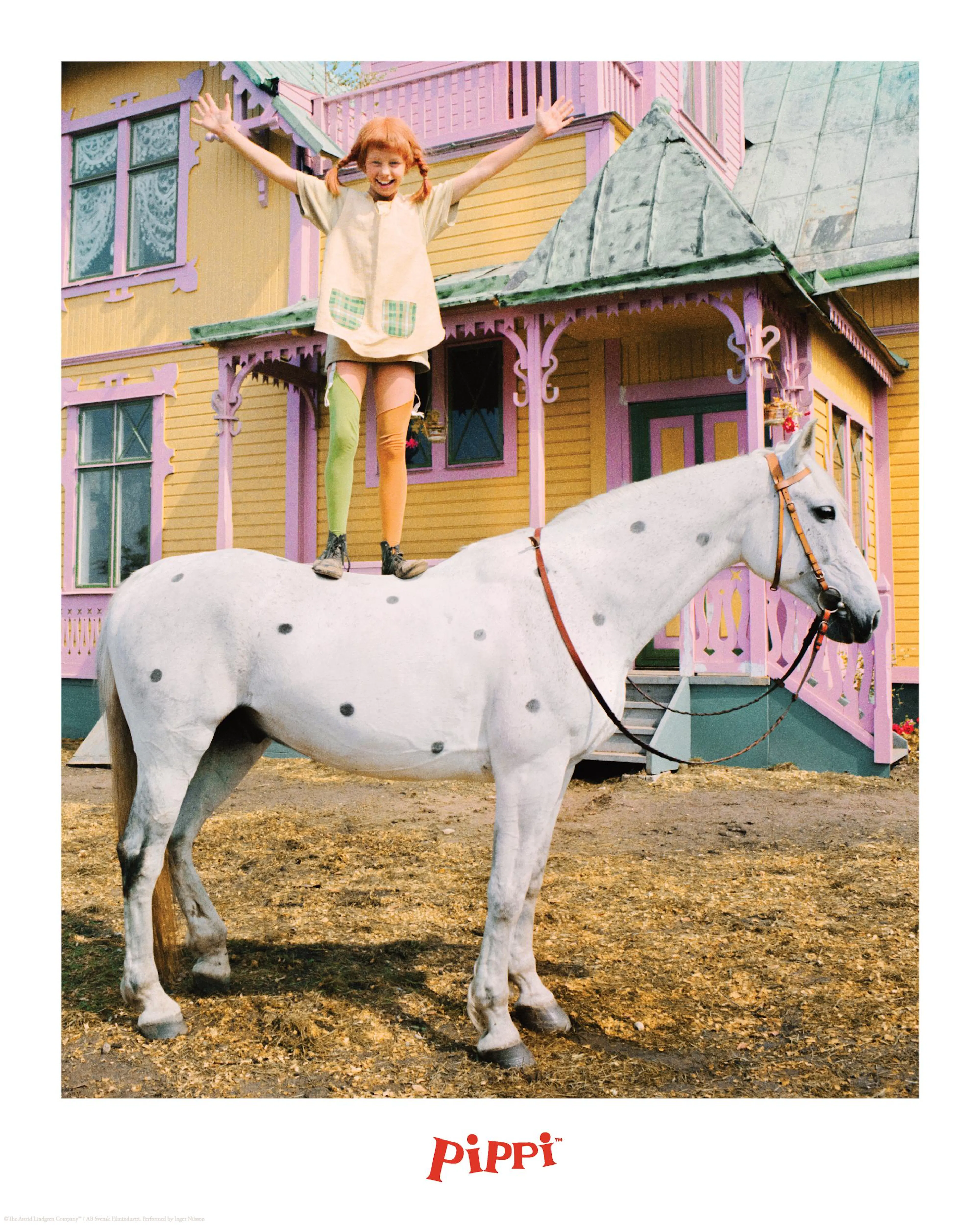 Pippi Longstocking standing on the white horse Little Old Man, arms outstretched, in front of a yellow and pink house.