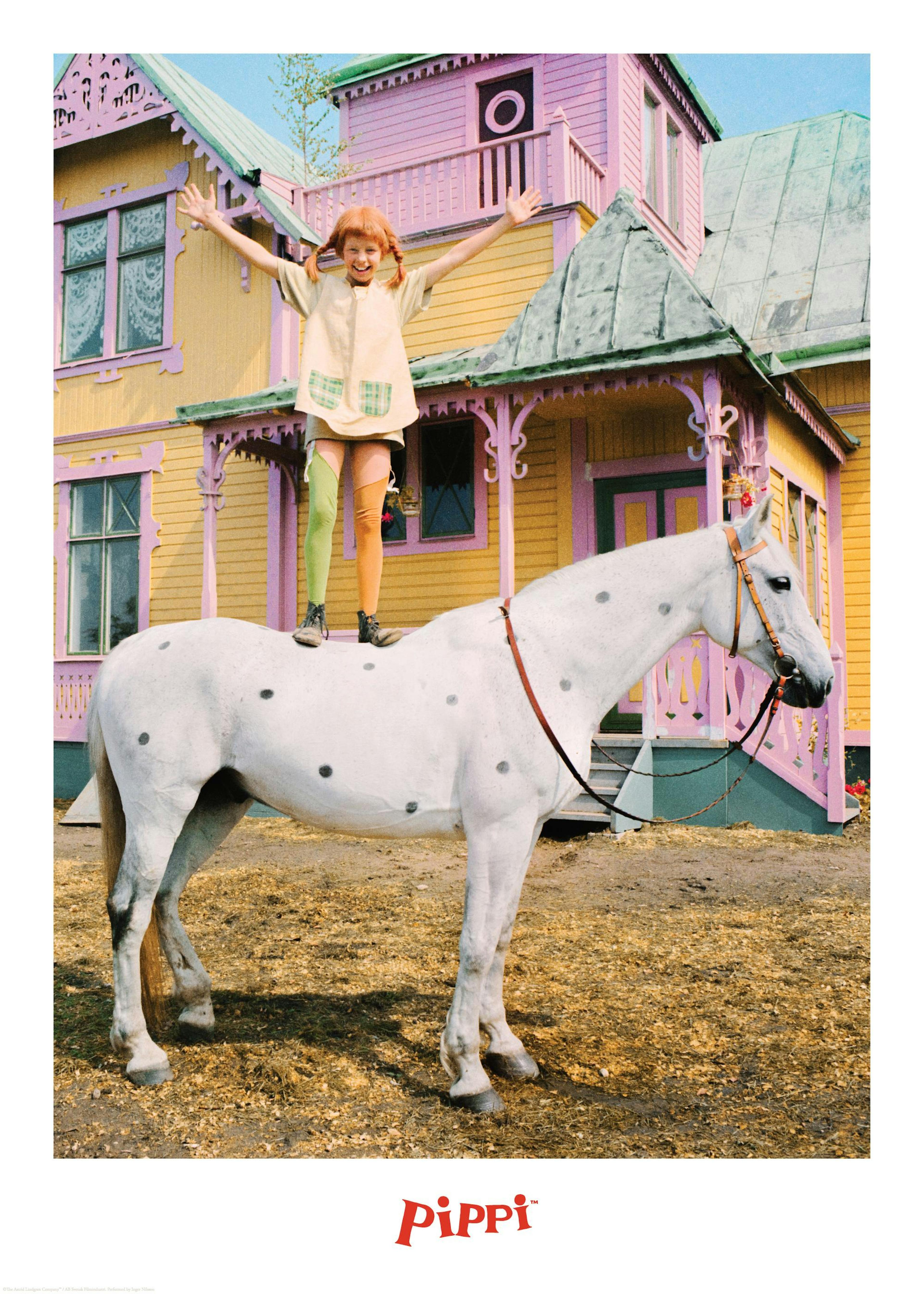 Pippi Langkous staat op Kleine Witje, haar witte paard met zwarte stippen, voor een geel huis met roze details. Pippi spreidt ha