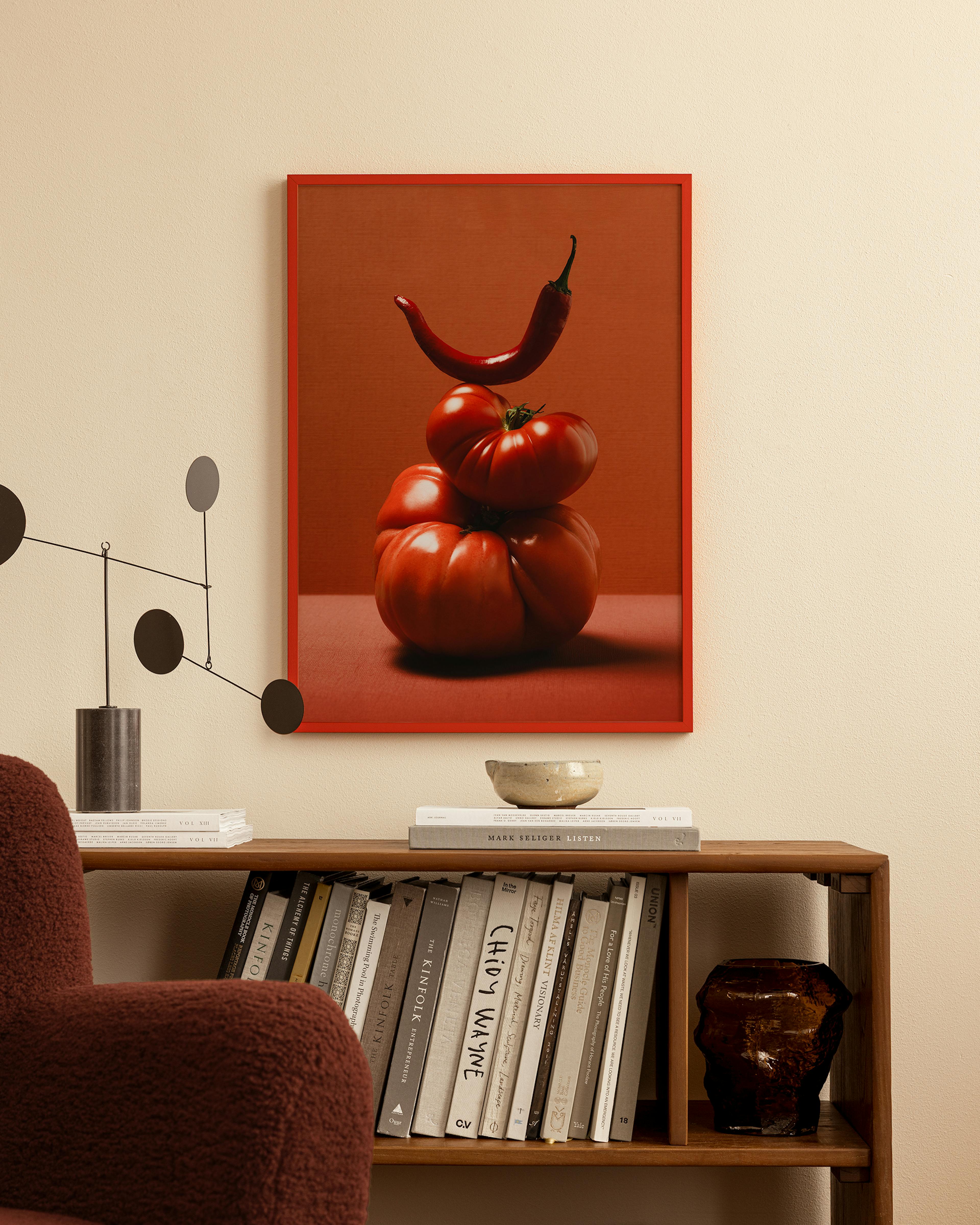 A print of stacked red tomatoes with a red chili on top, displayed above a wooden bookshelf in a living room.