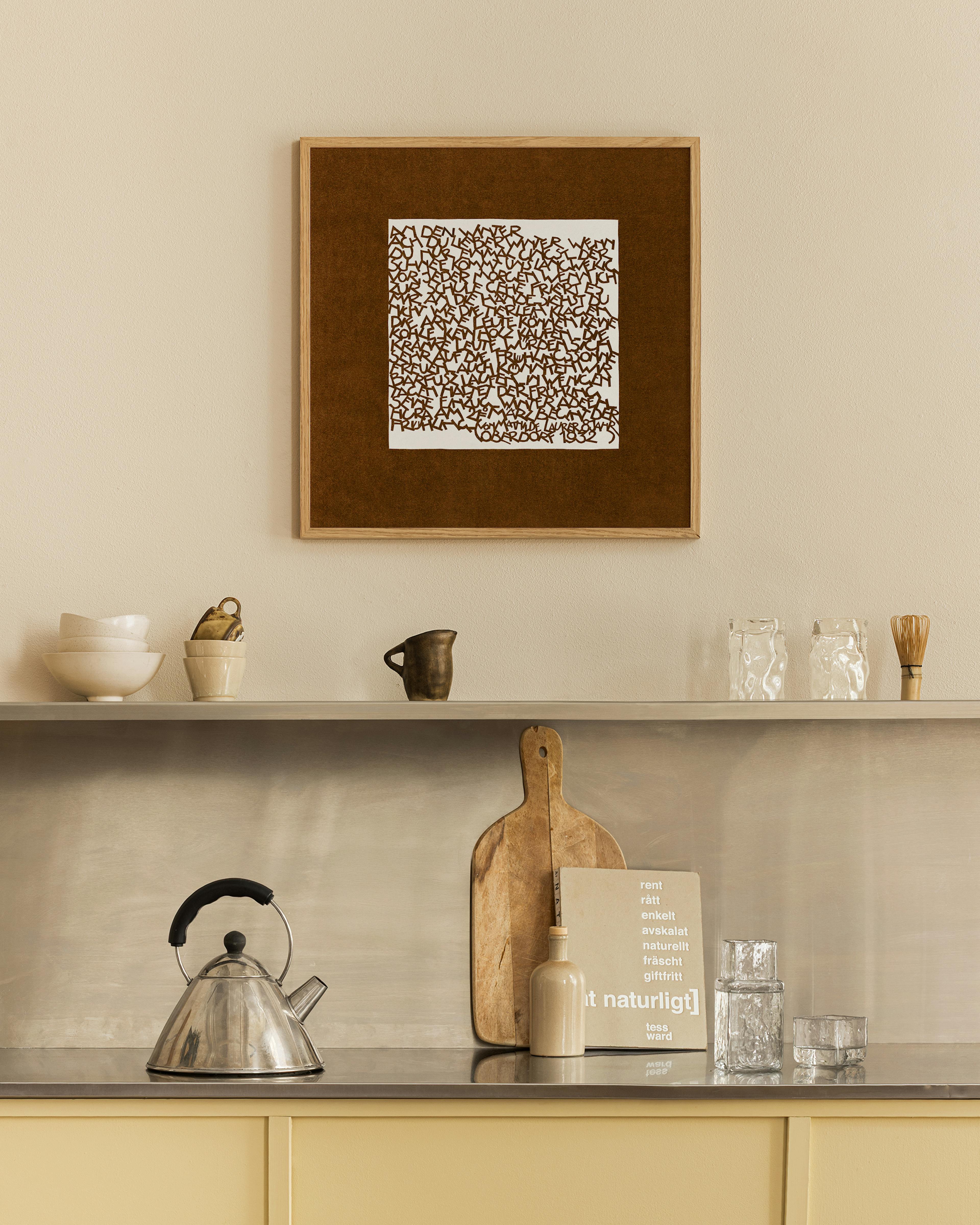 Póster de arte tipográfico con letras marrones sobre fondo blanco y marrón, colgado sobre una repisa en una cocina moderna.