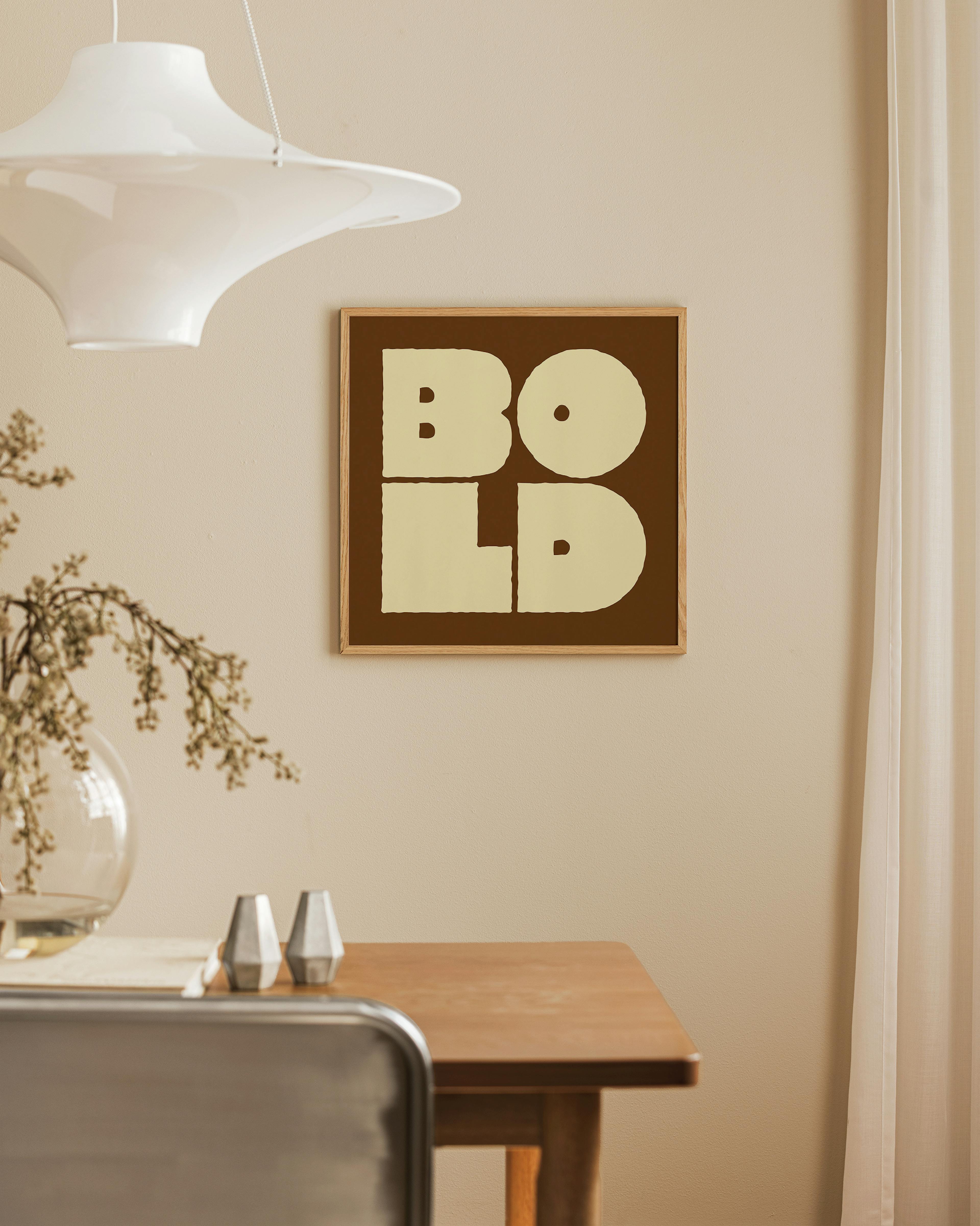 Póster BOLD en letras crema sobre fondo marrón, colgando en una pared beige sobre una mesa de madera con un jarrón.