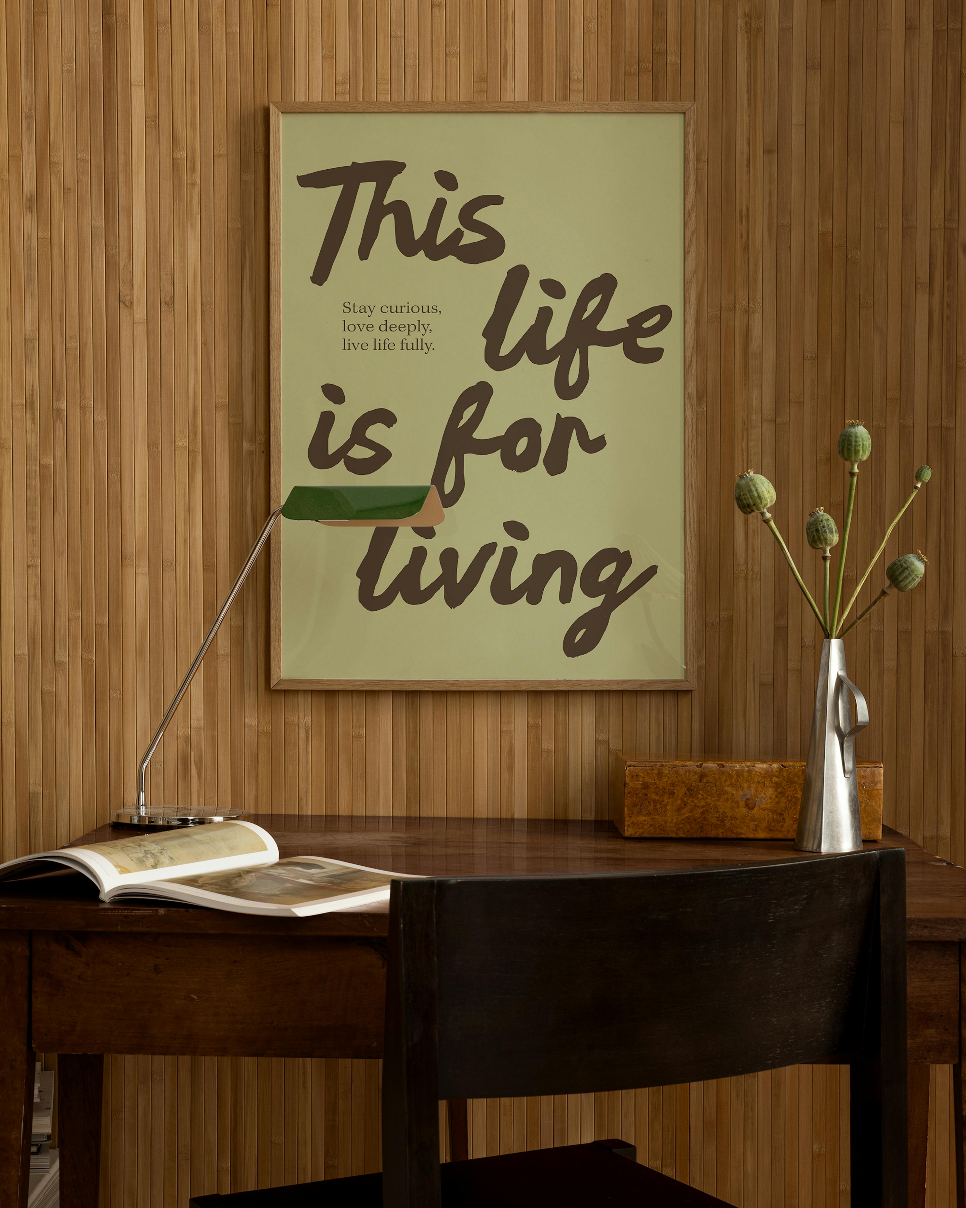 Póster This life is for living en verde claro y letras marrones, colgado sobre un escritorio de madera con una lámpara y un jarr