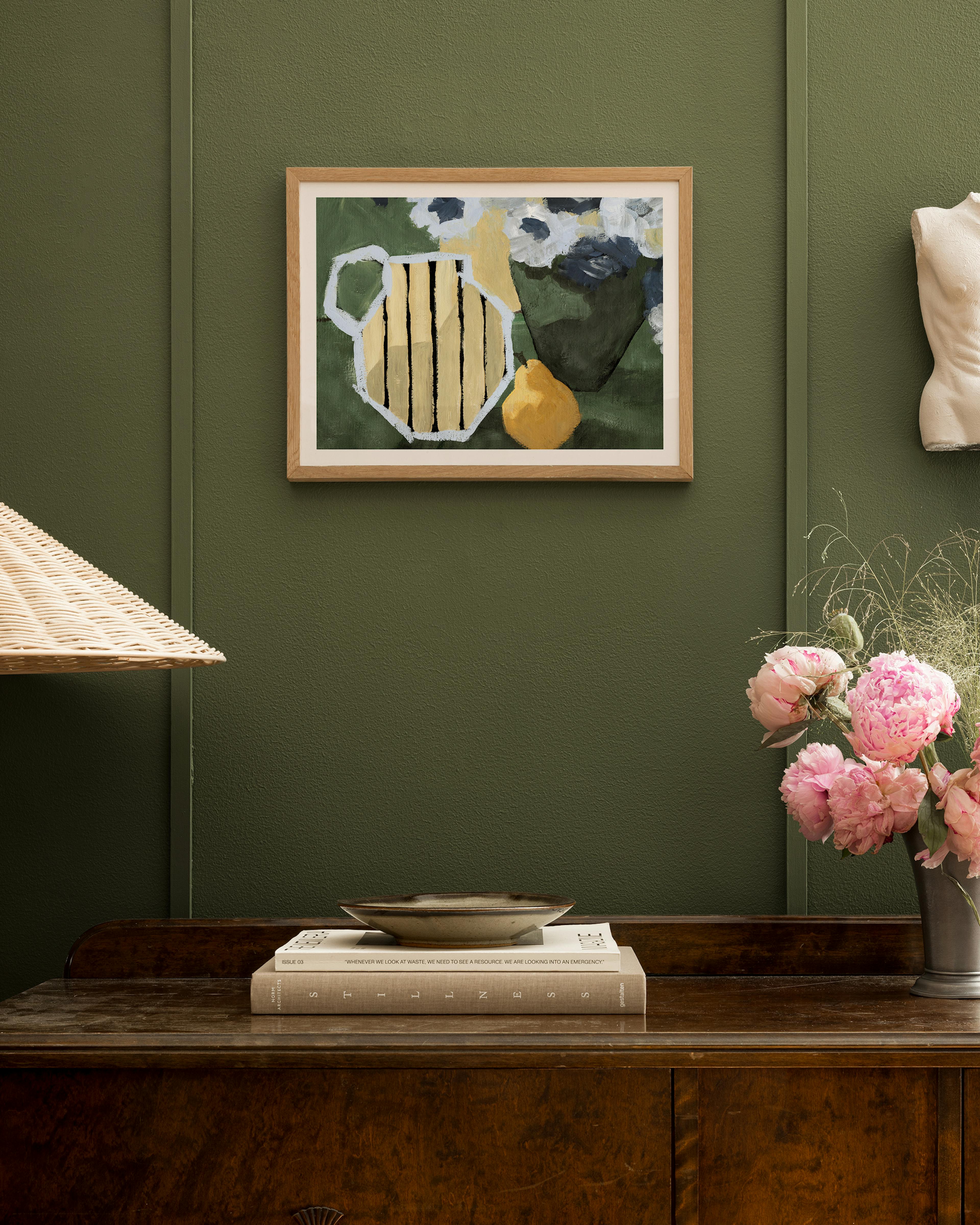 Still life print with a yellow pitcher and pear, and white abstract flowers, hanging in a dark green room above a wooden dresser