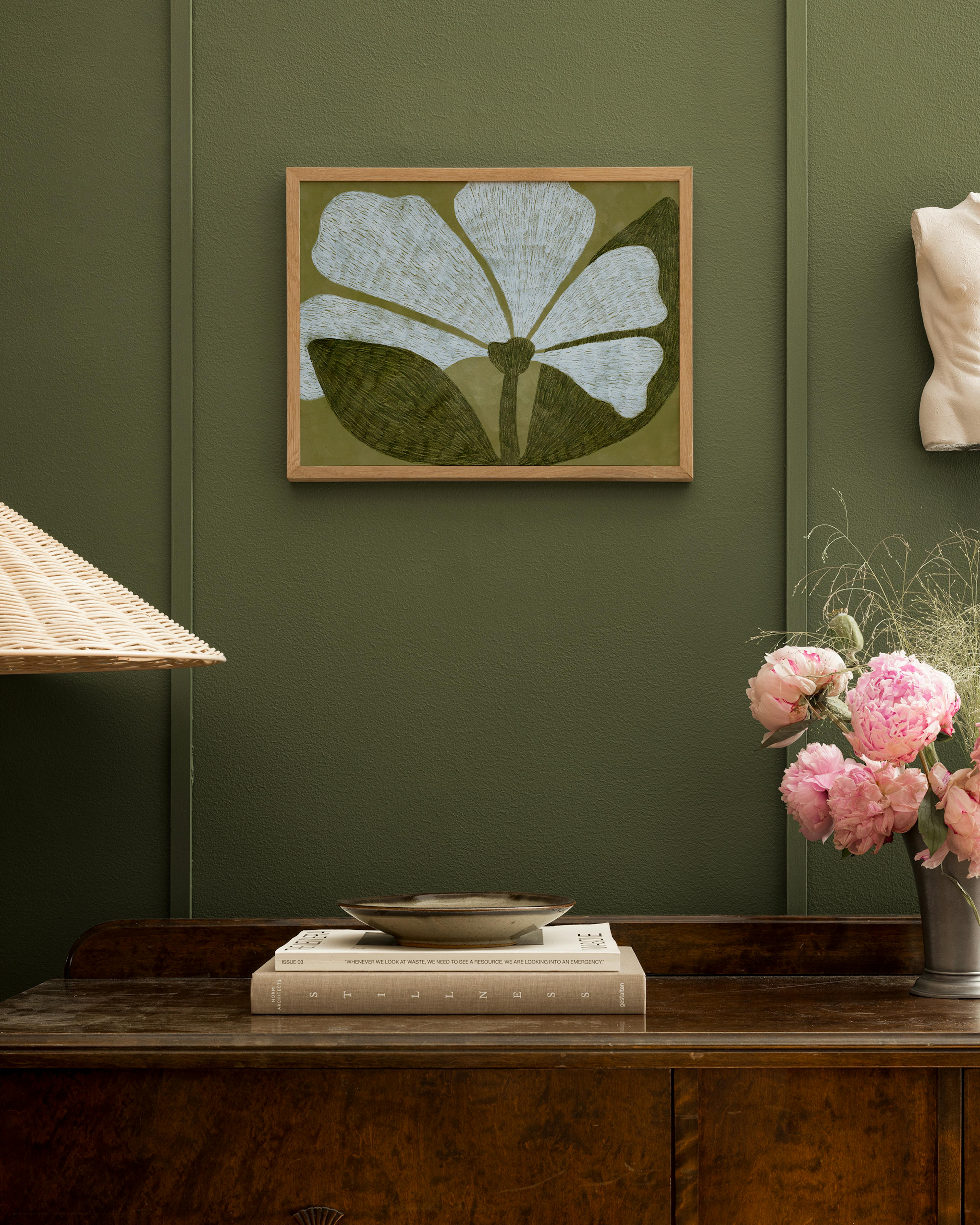 Póster de una flor de pétalos blancos con hojas verdes oscuras enmarcado, colgado en una pared verde oliva encima de un mueble d
