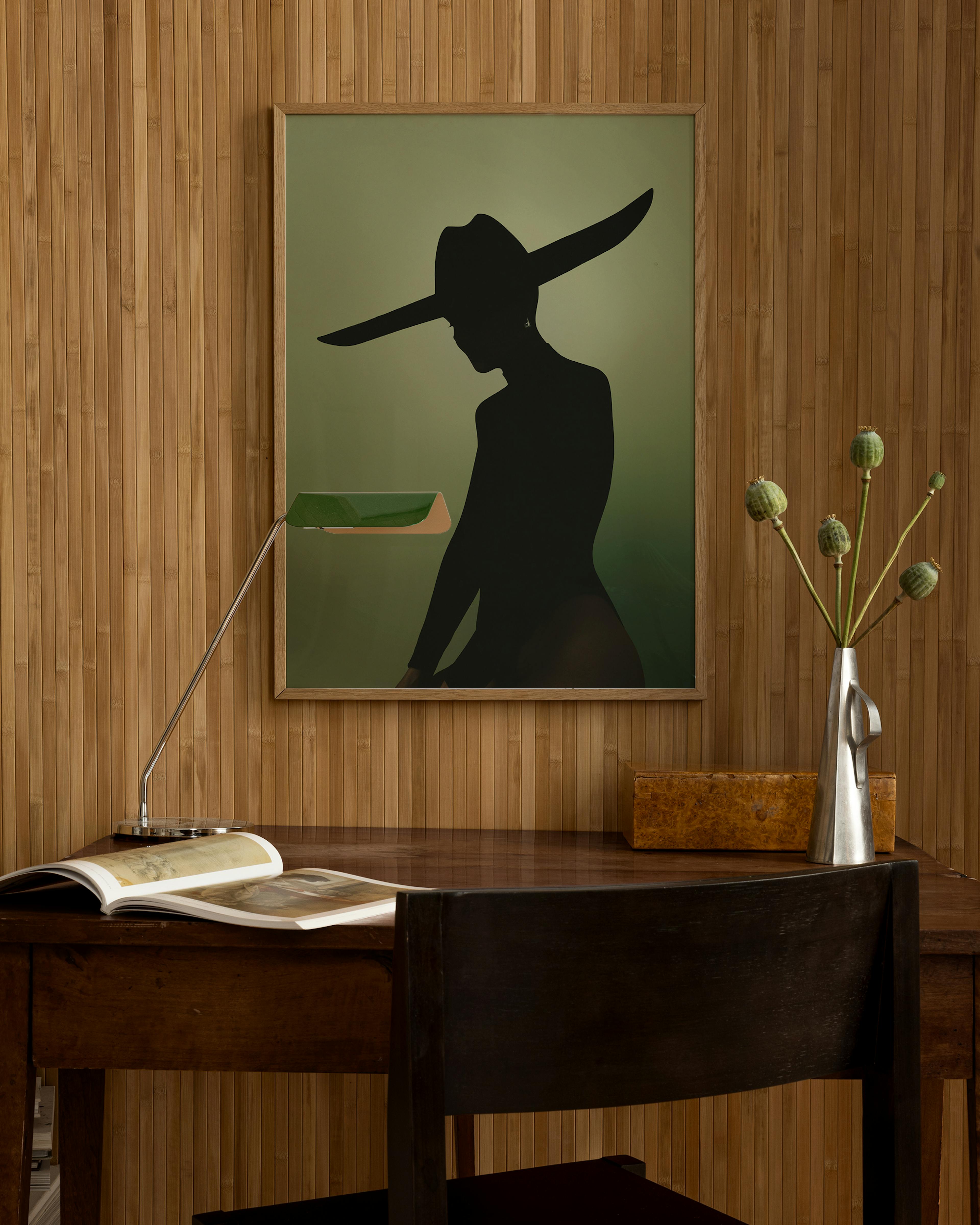 A black silhouette of a woman in a wide-brimmed hat on a green background, hanging above a wooden desk.