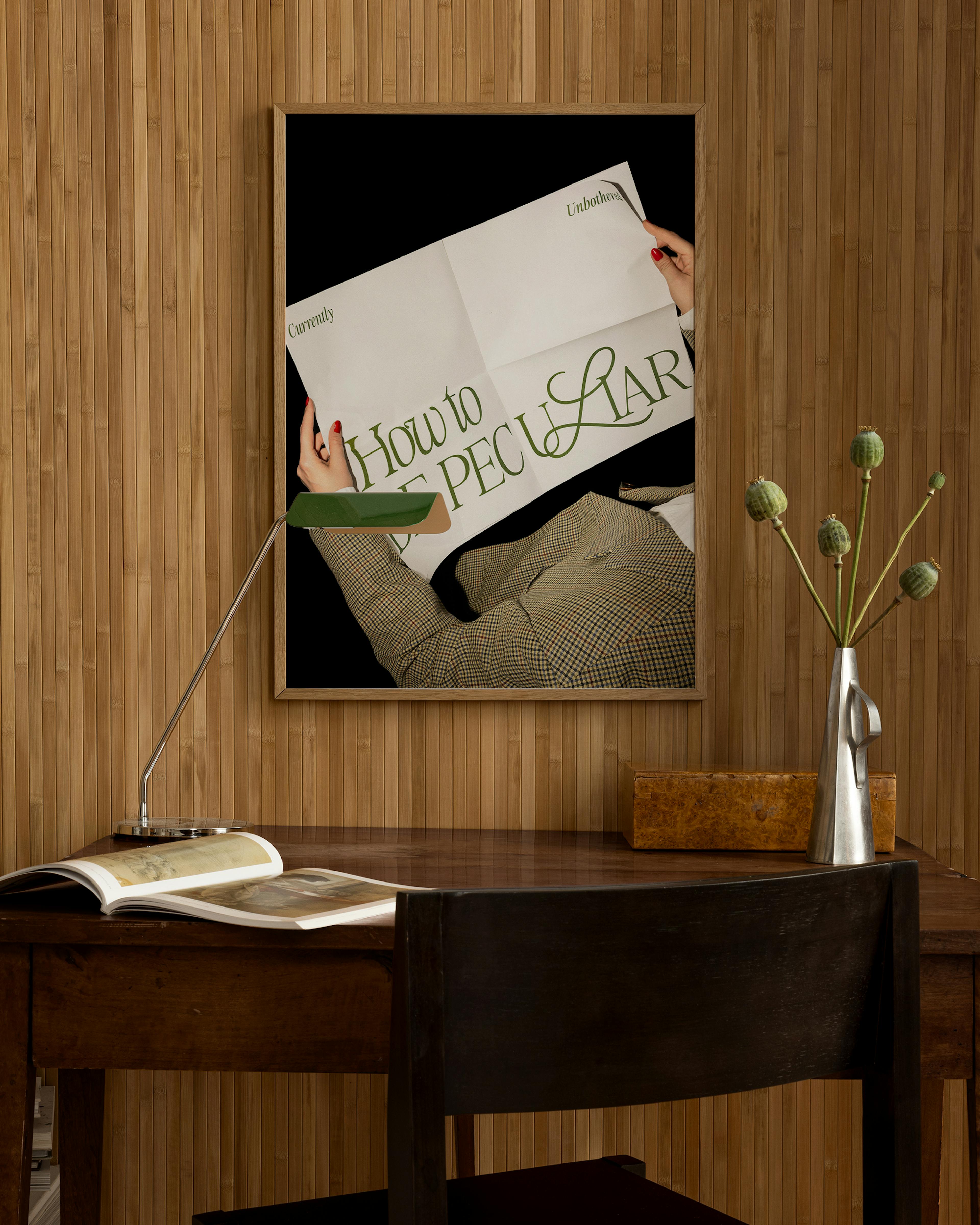 A print of a person in a plaid jacket holding a newspaper with How to be Peculiar in green, hanging above a wooden desk with a l