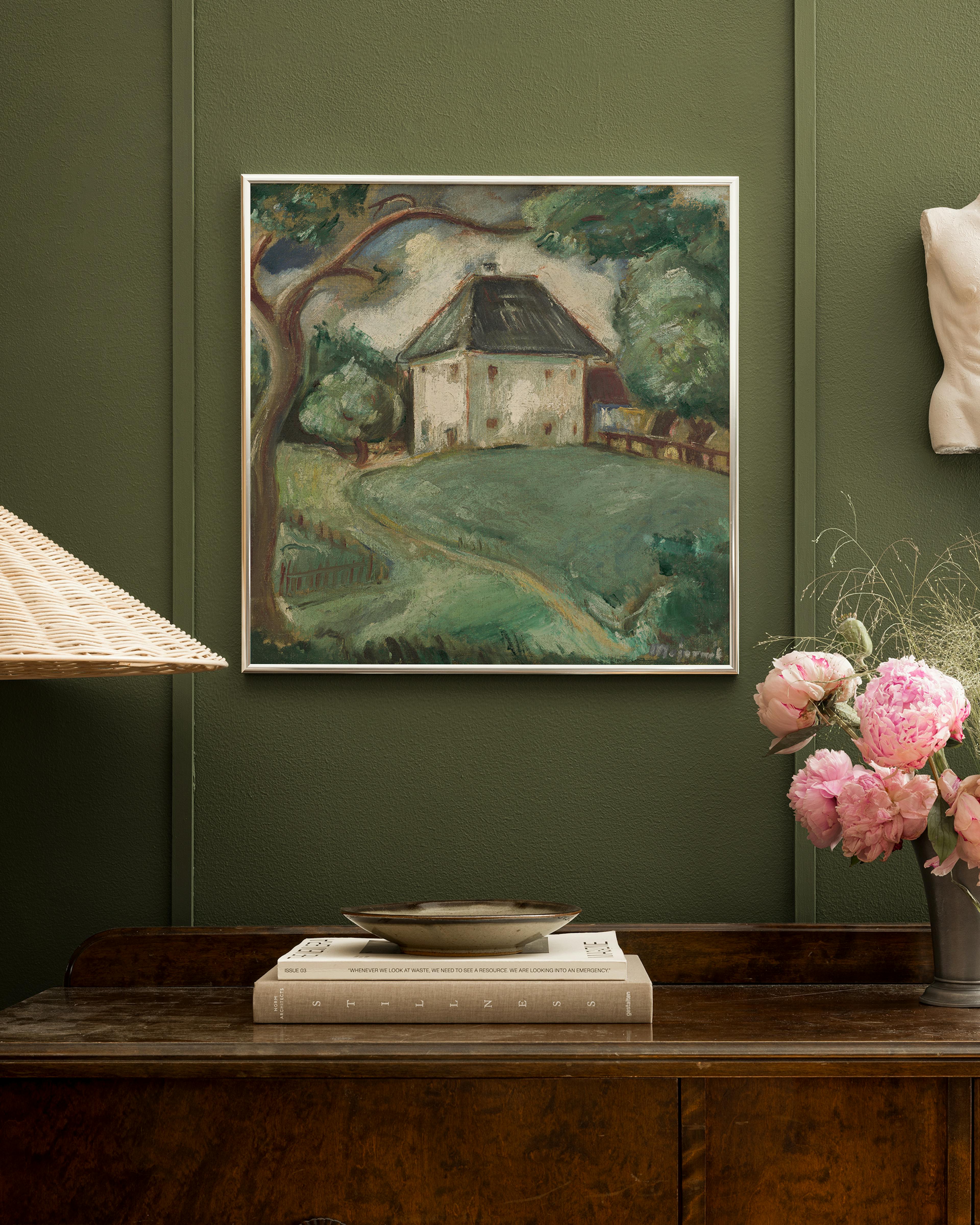 A landscape print of a white house with a dark roof and green grounds, hanging on a dark green wall above a wooden chest with bo