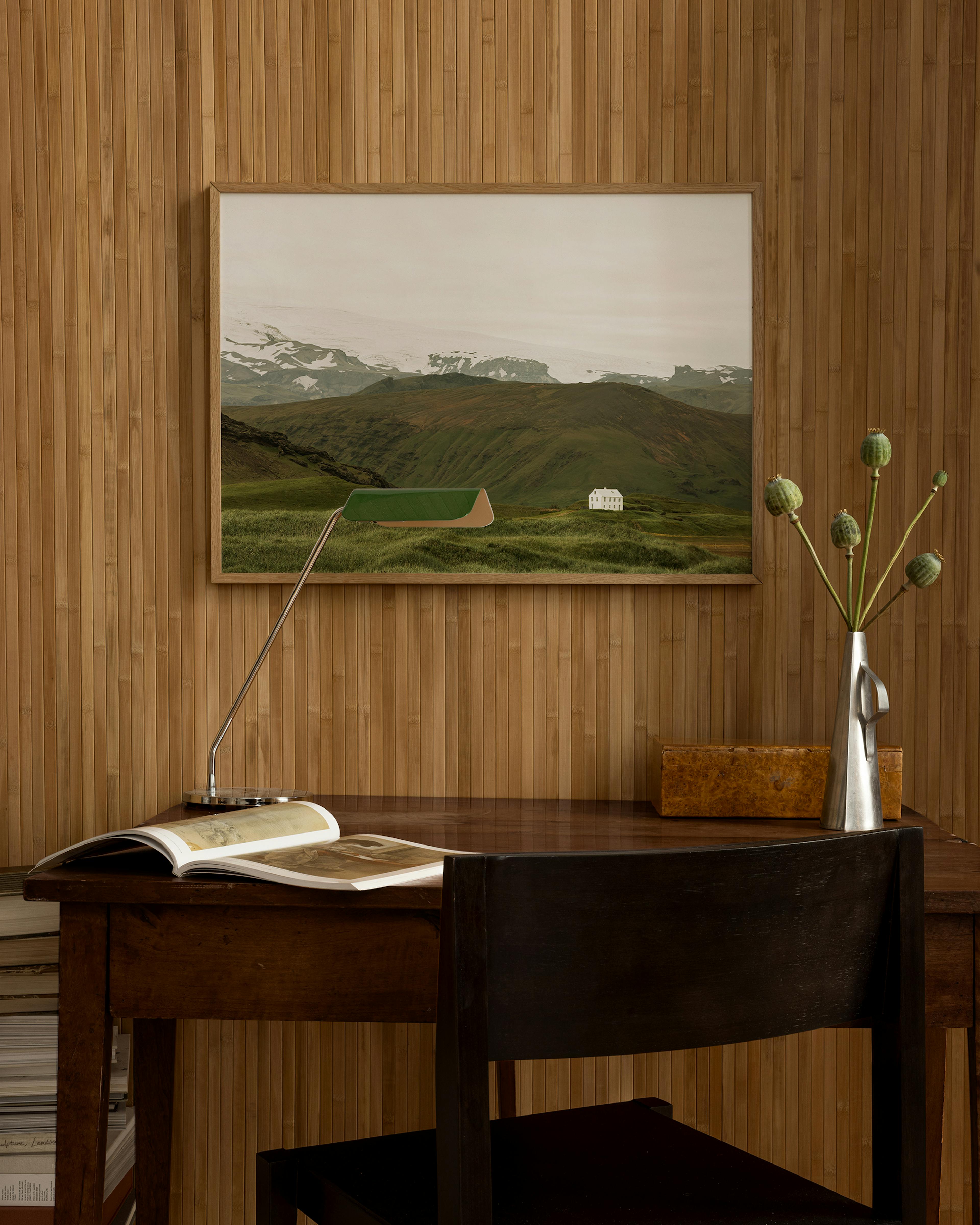 A landscape print of a white house in a green valley with snow-capped mountains, displayed above a dark wood desk.