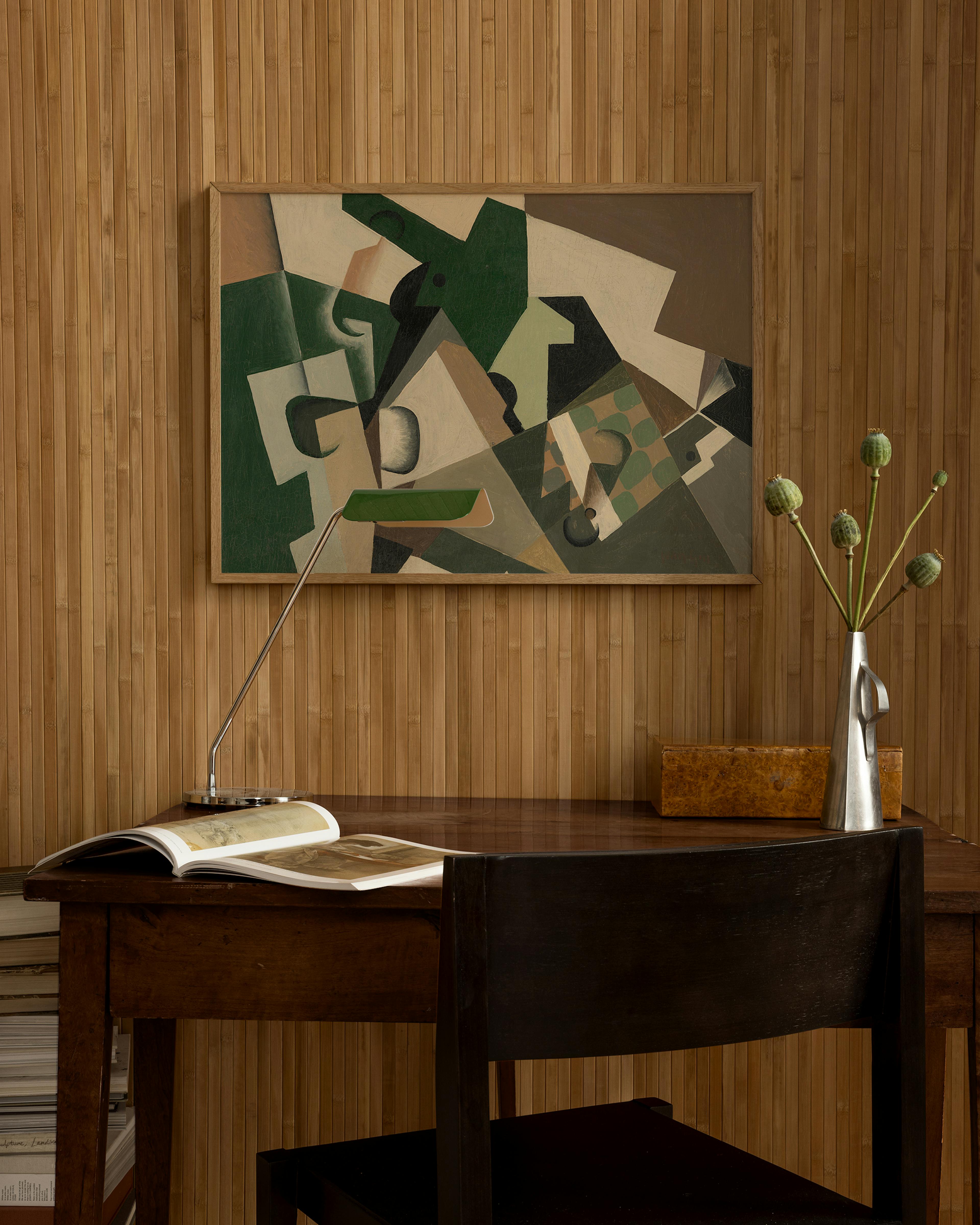 Cubist print with green, brown, and off-white fragmented shapes, hanging above a wooden desk with a lamp, book, and vase.