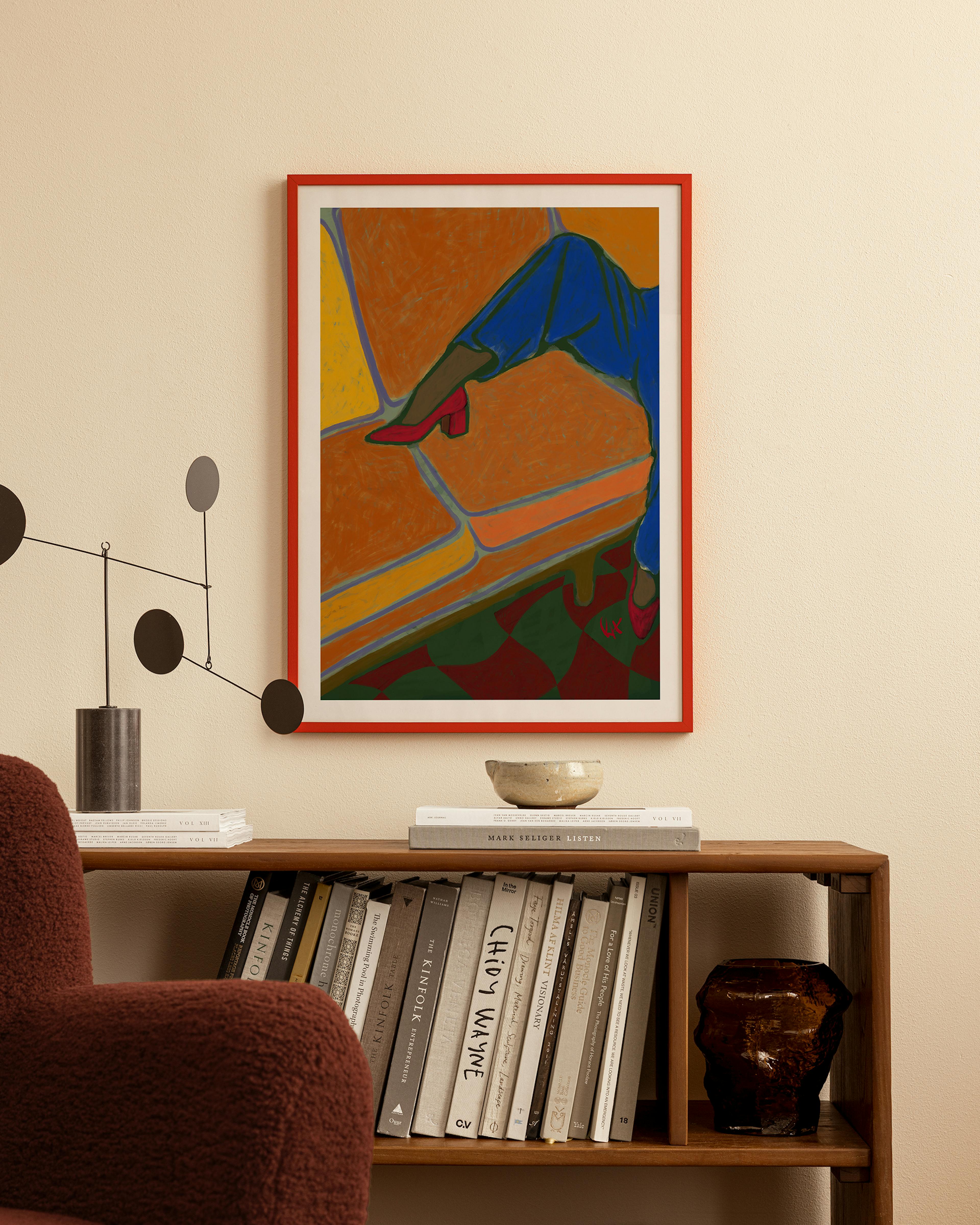 A colorful print of a persons leg in blue pants and red heels, resting on an orange sofa, hanging above a wooden bookshelf.