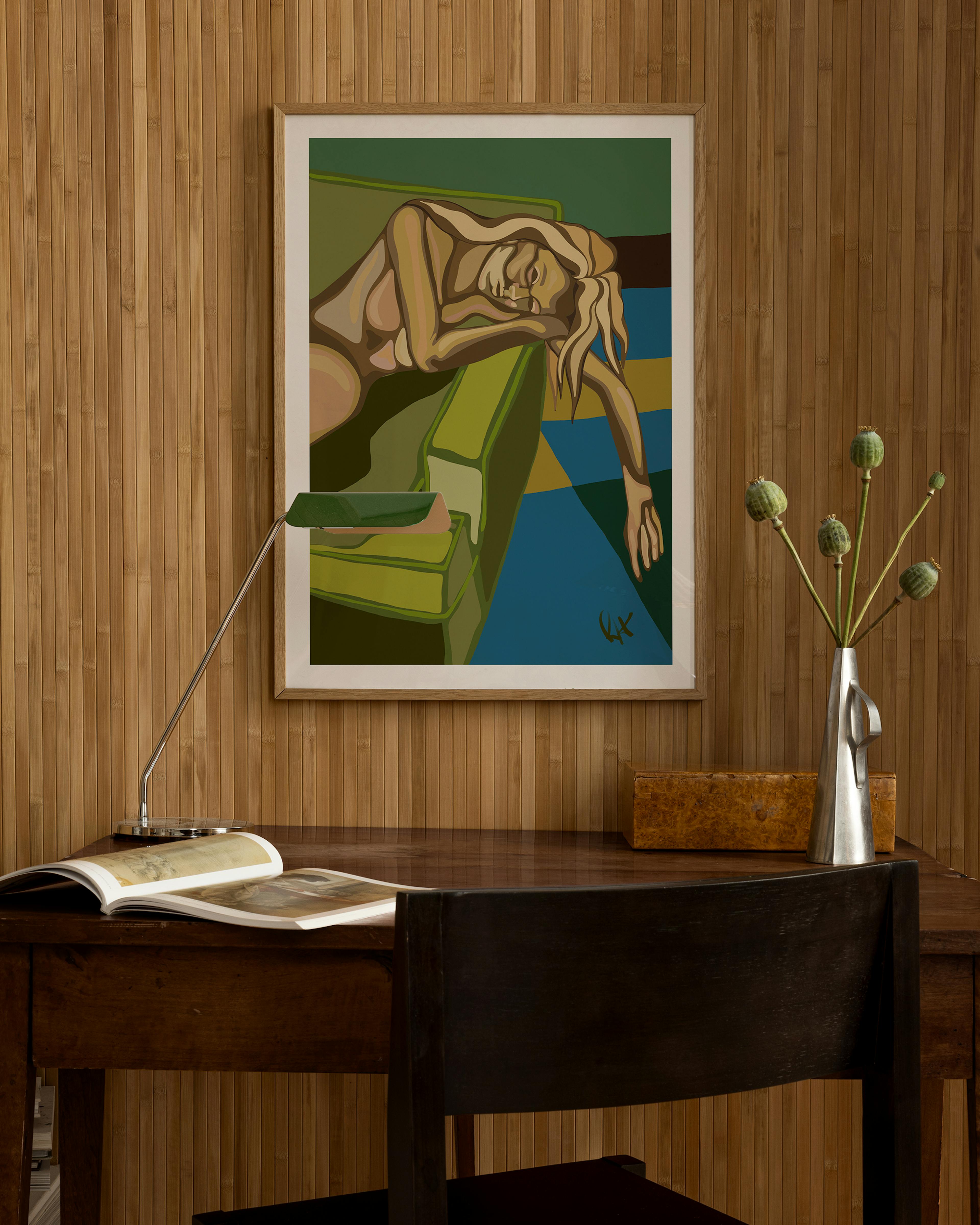 A colorful print of a sleeping woman on a green couch, hanging above a wooden desk with a lamp, book, and vase.
