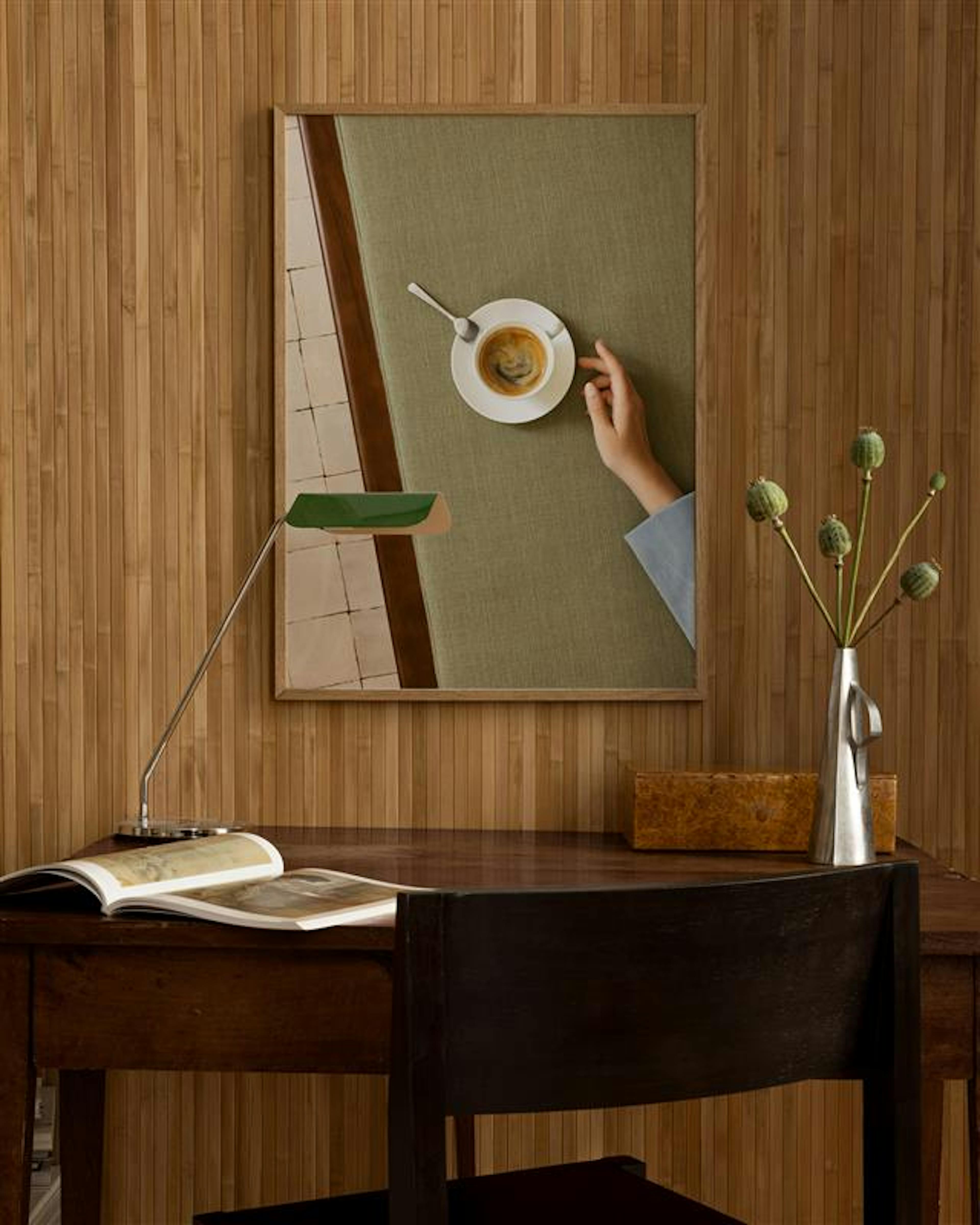 Un póster de café mostrando una mano y una taza, colgado sobre un escritorio de madera con una lámpara y un jarrón.