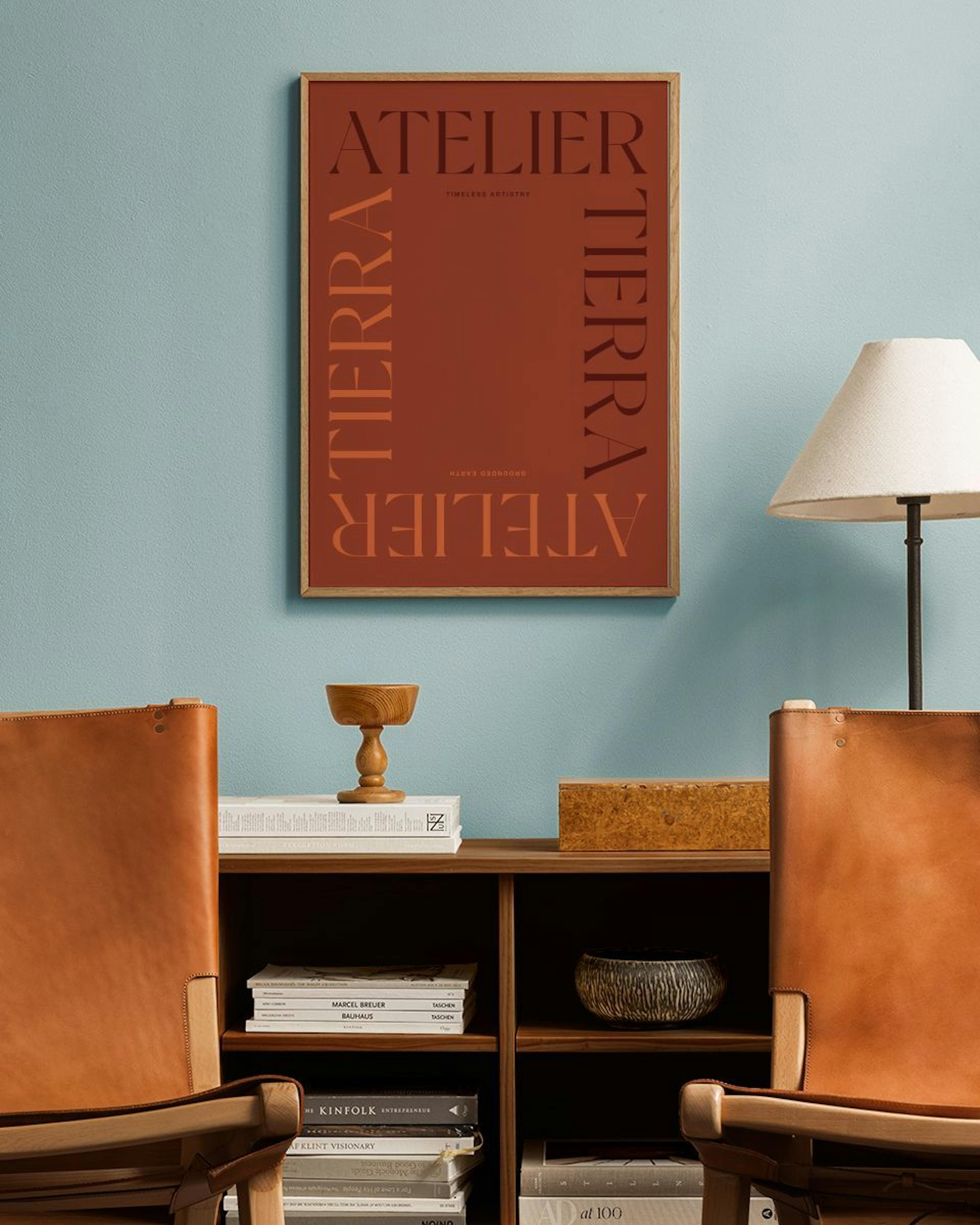 Póster Atelier Tierra en tonos tierra sobre una pared azul, en una sala con sillas de cuero y estantería de madera.