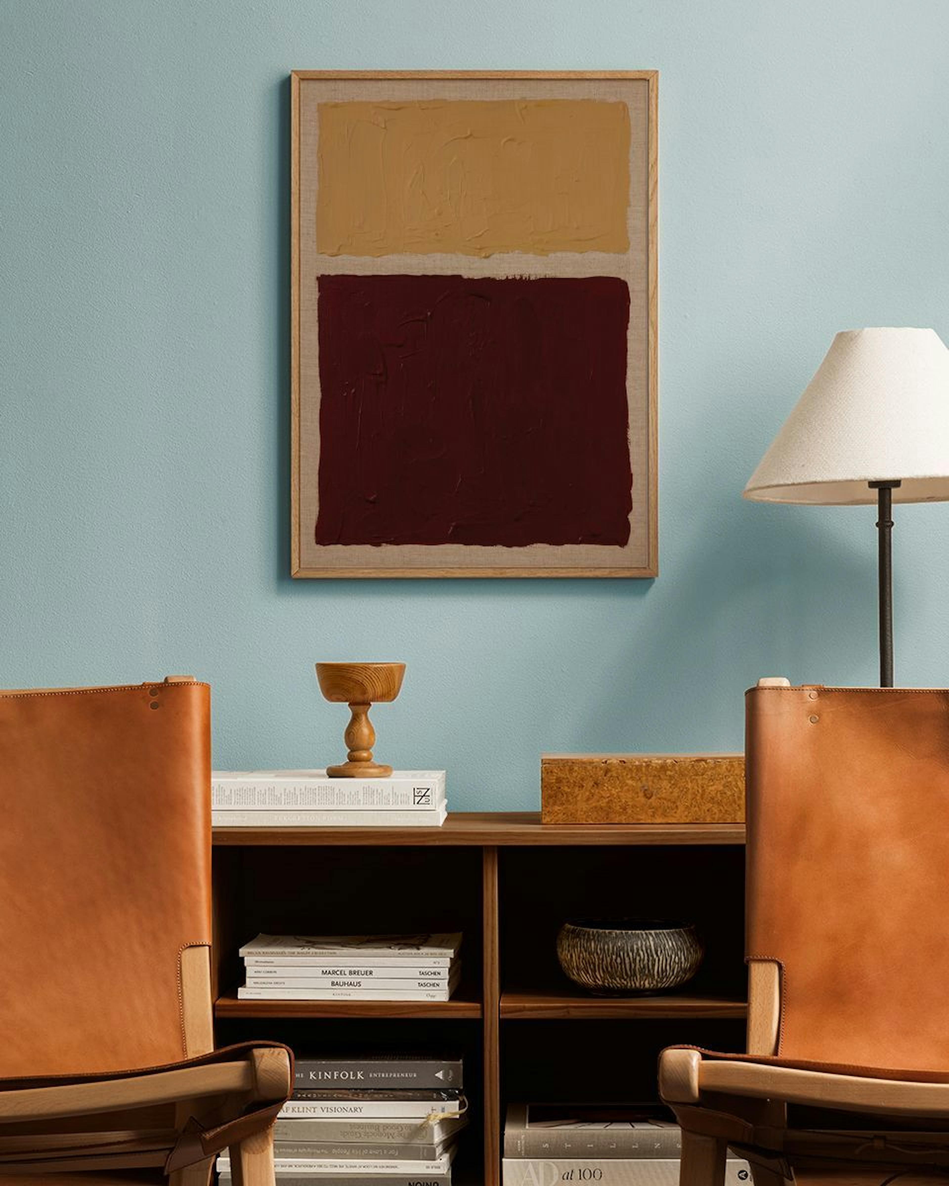 A print of two textured blocks of color, one ochre and one deep red, hanging above a wooden bookshelf between two leather chairs