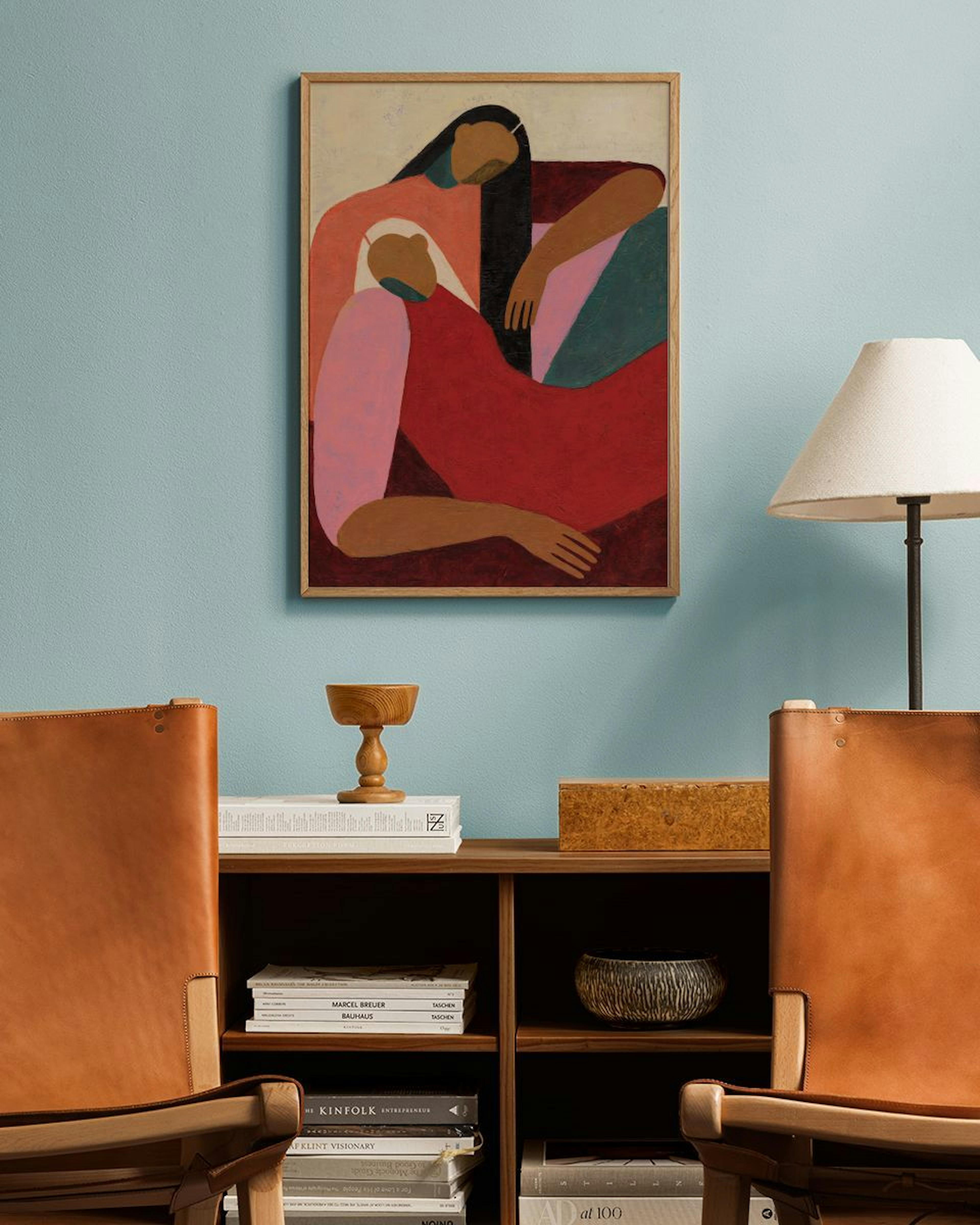 Abstract print of two figures in warm tones of red, pink, and green, hanging on a light blue wall above a wooden shelf.