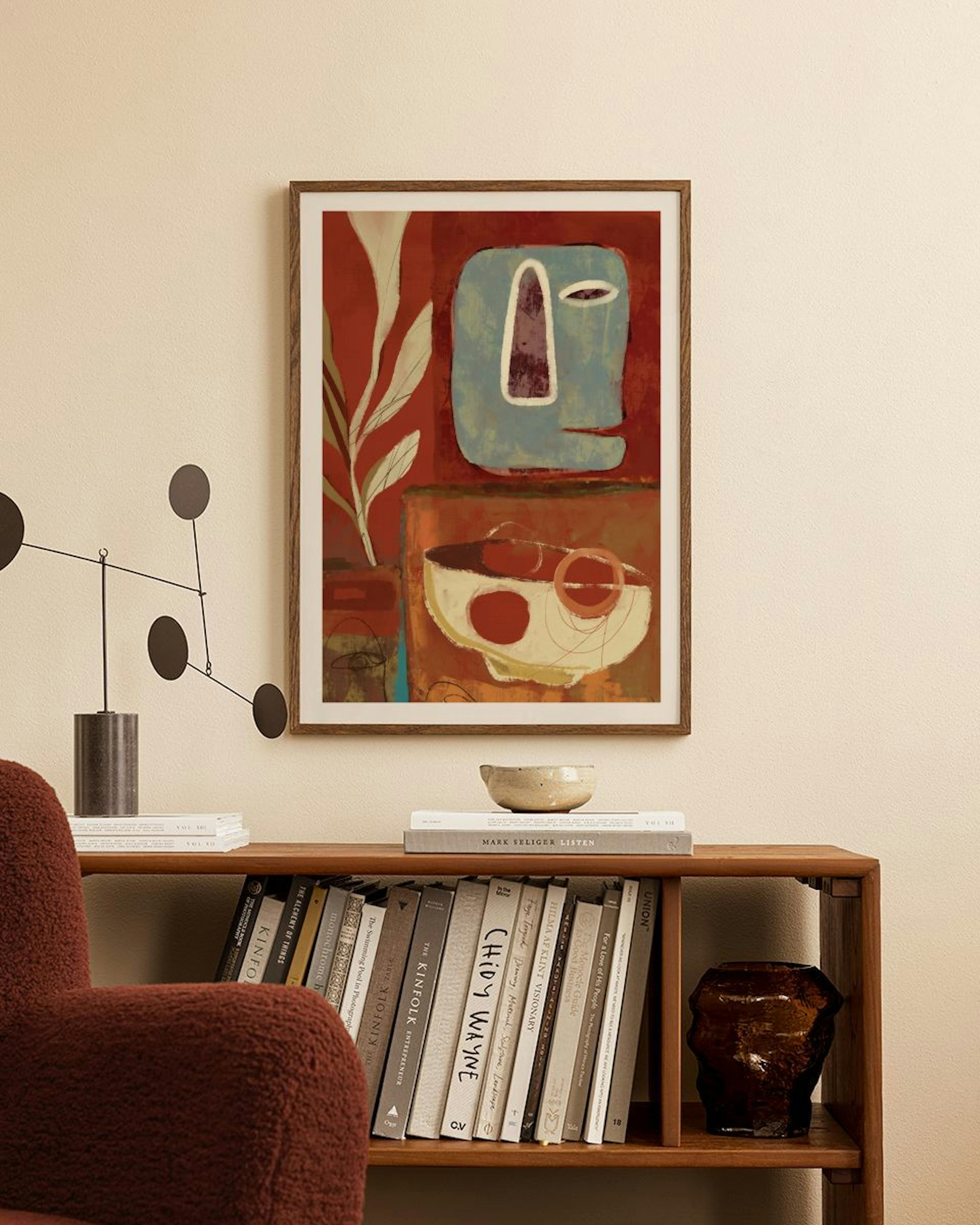 Póster abstracto con rostro azul, planta y tazón en tonos rojos, colgando sobre un estante de madera con libros.