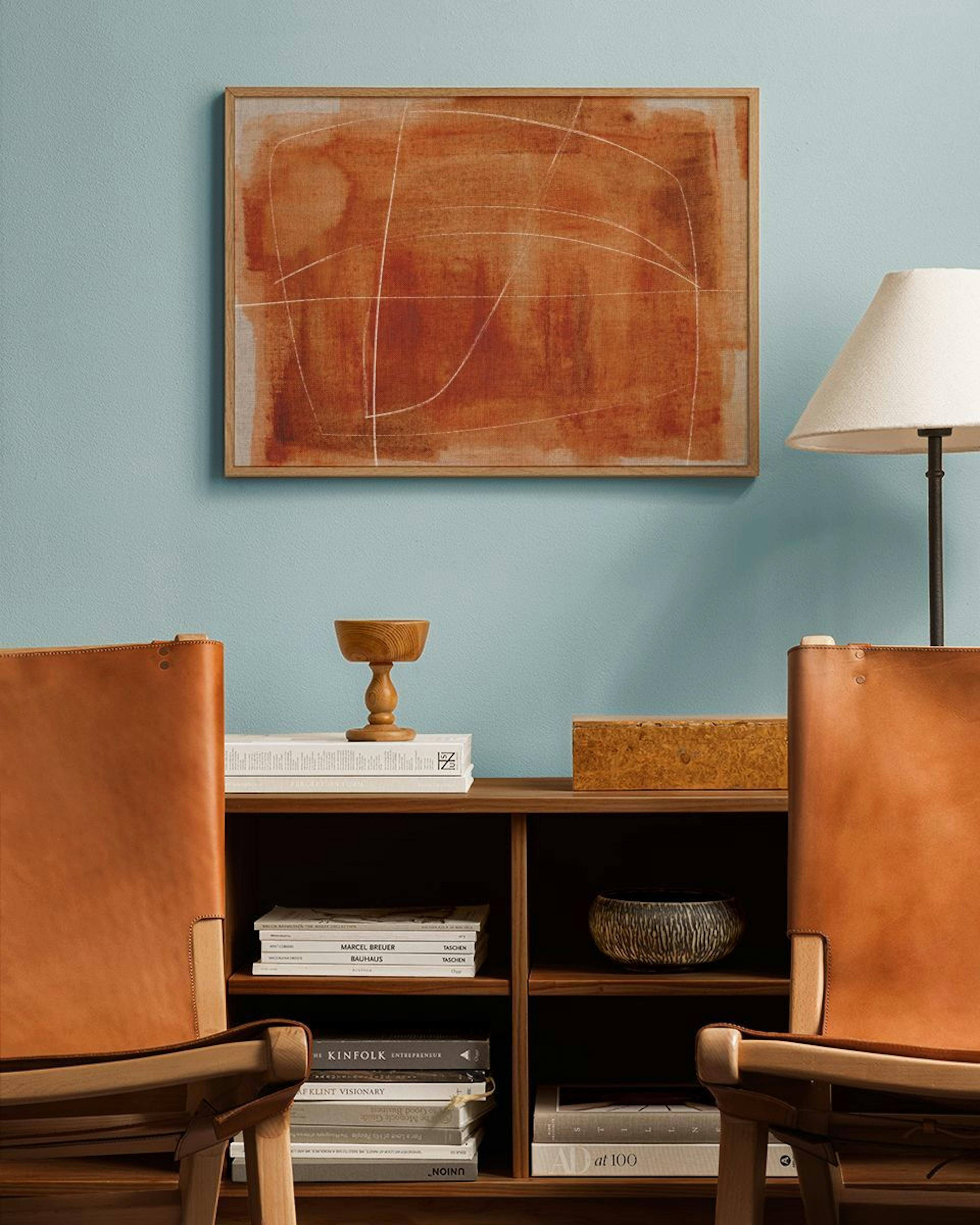 An abstract print with rust-colored brushstrokes and flowing white lines, hanging above a wooden bookshelf with books and decor 