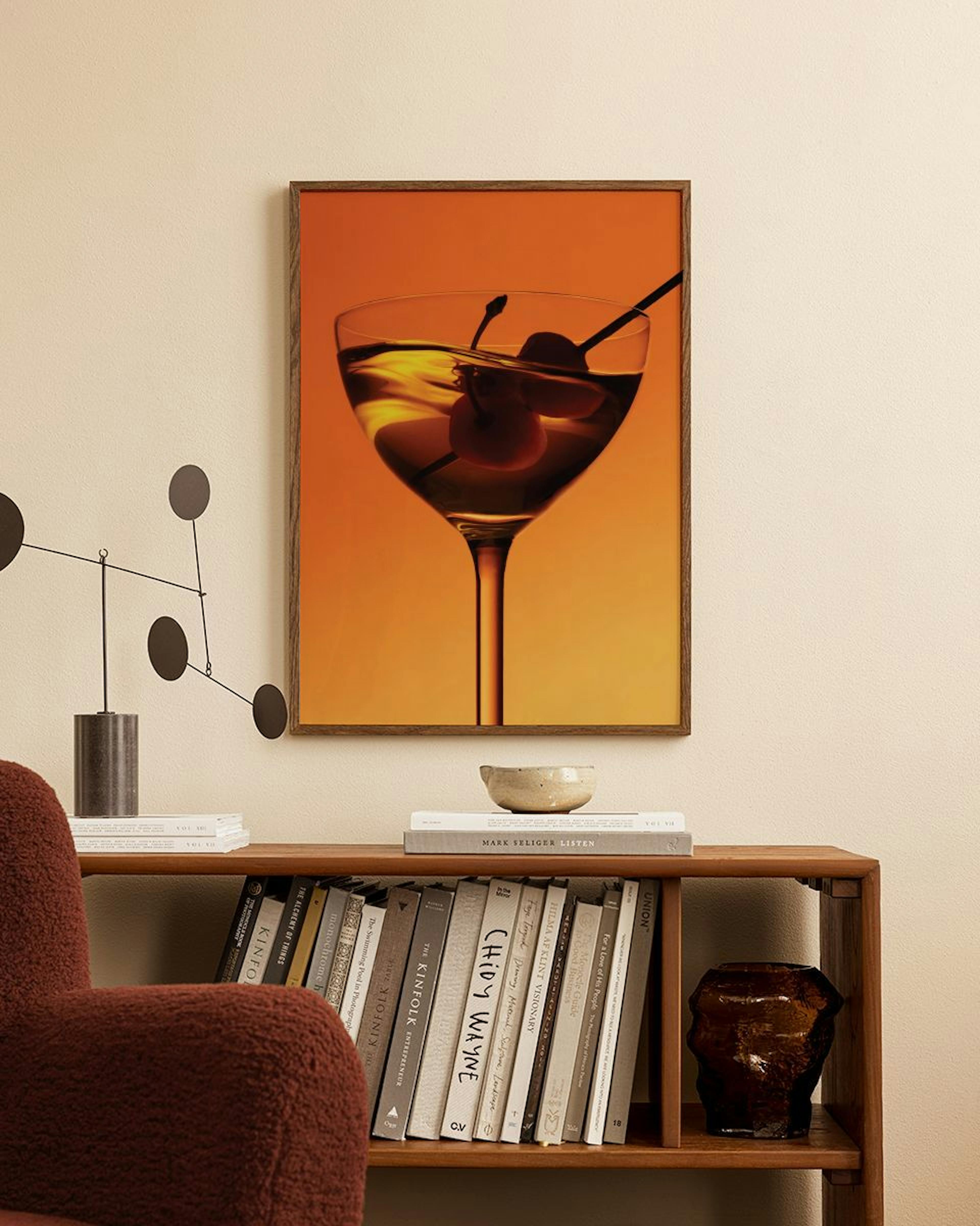 A print of a martini glass with two cherries and a stirrer, set against a warm orange background, hanging above a wooden bookshe
