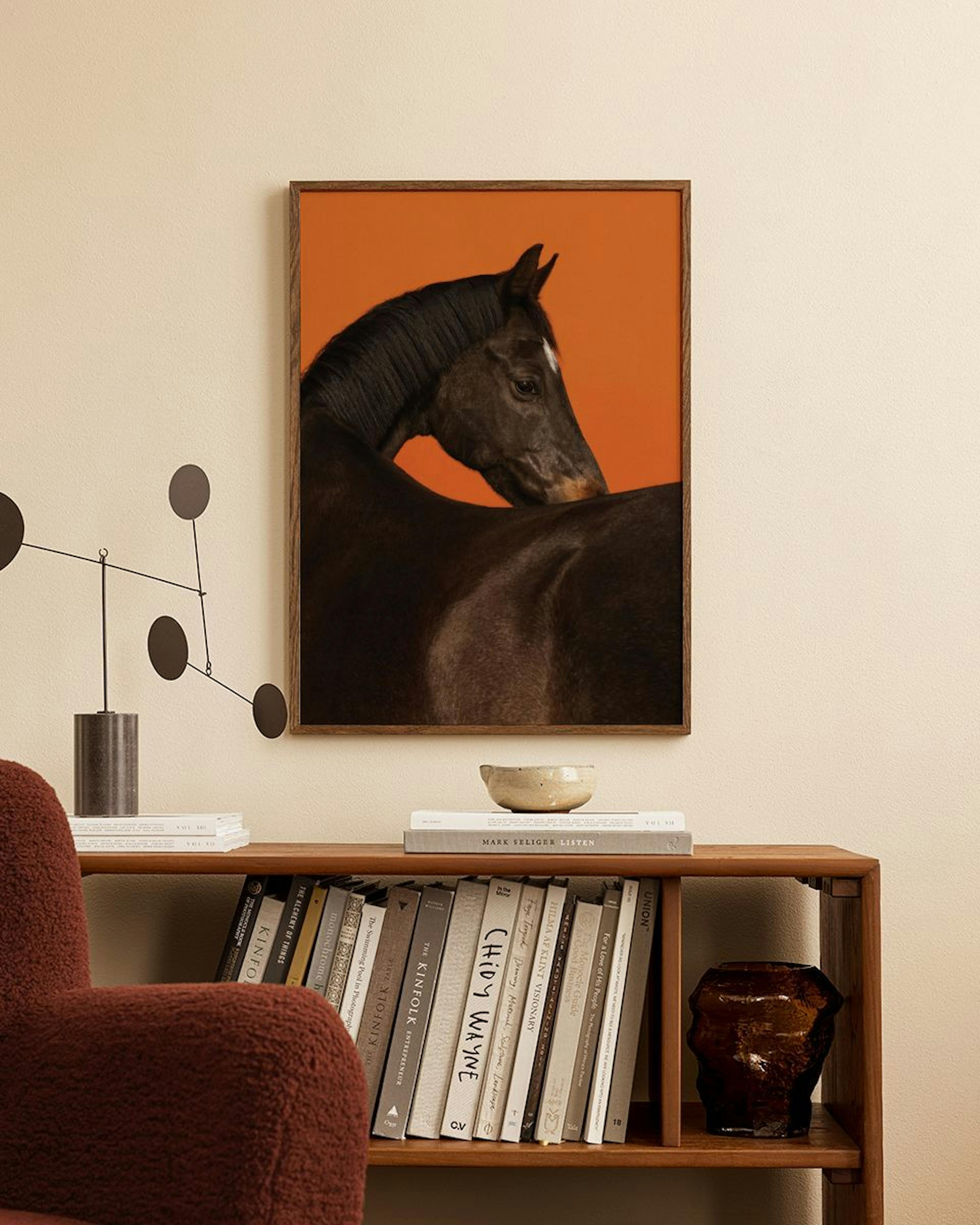 A print of a dark horse with a white blaze on an orange background, hanging above a wooden bookshelf in a cozy room.