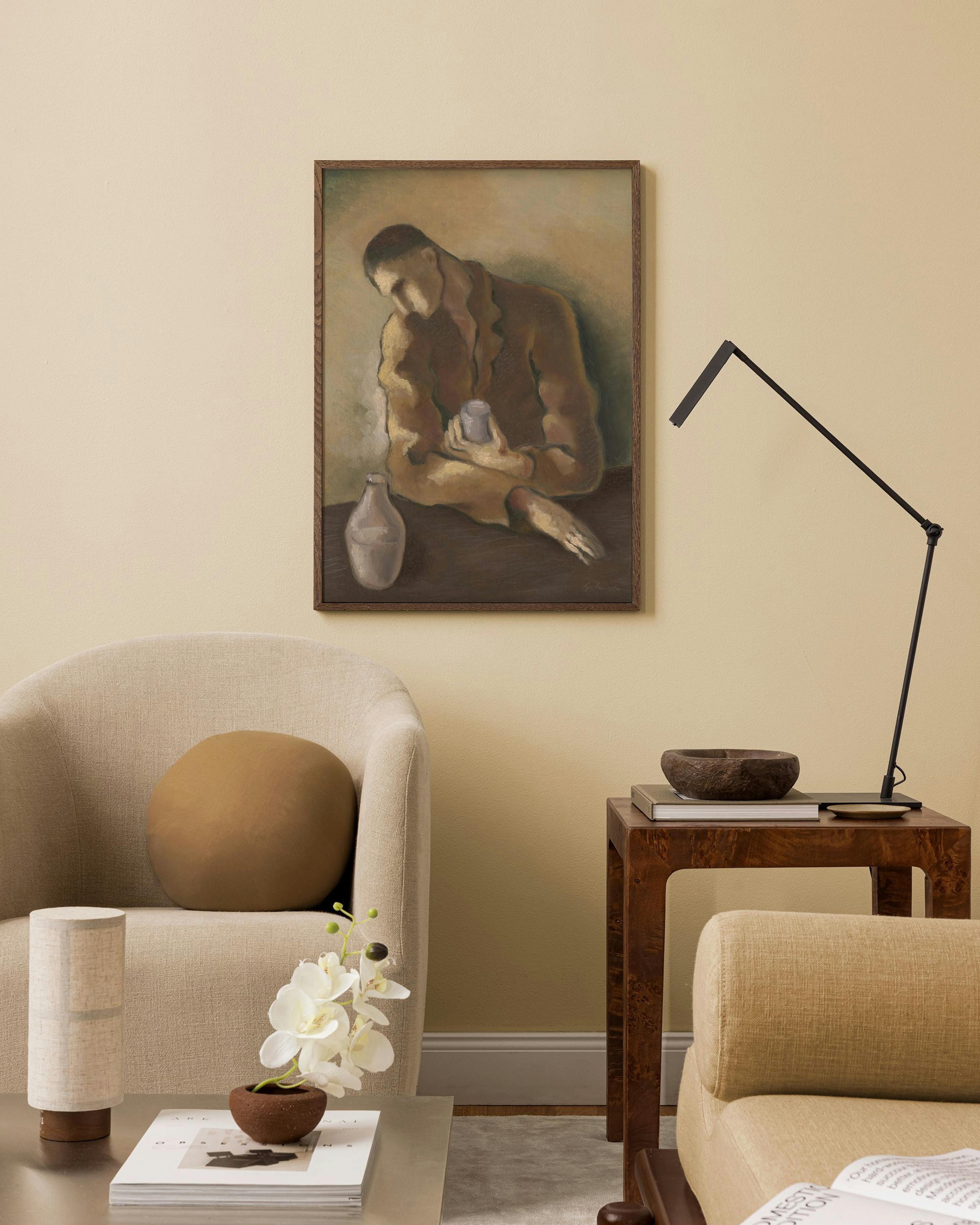 A print of a seated figure in brown tones with a jug, hanging in a neutral-toned living room above a beige armchair.