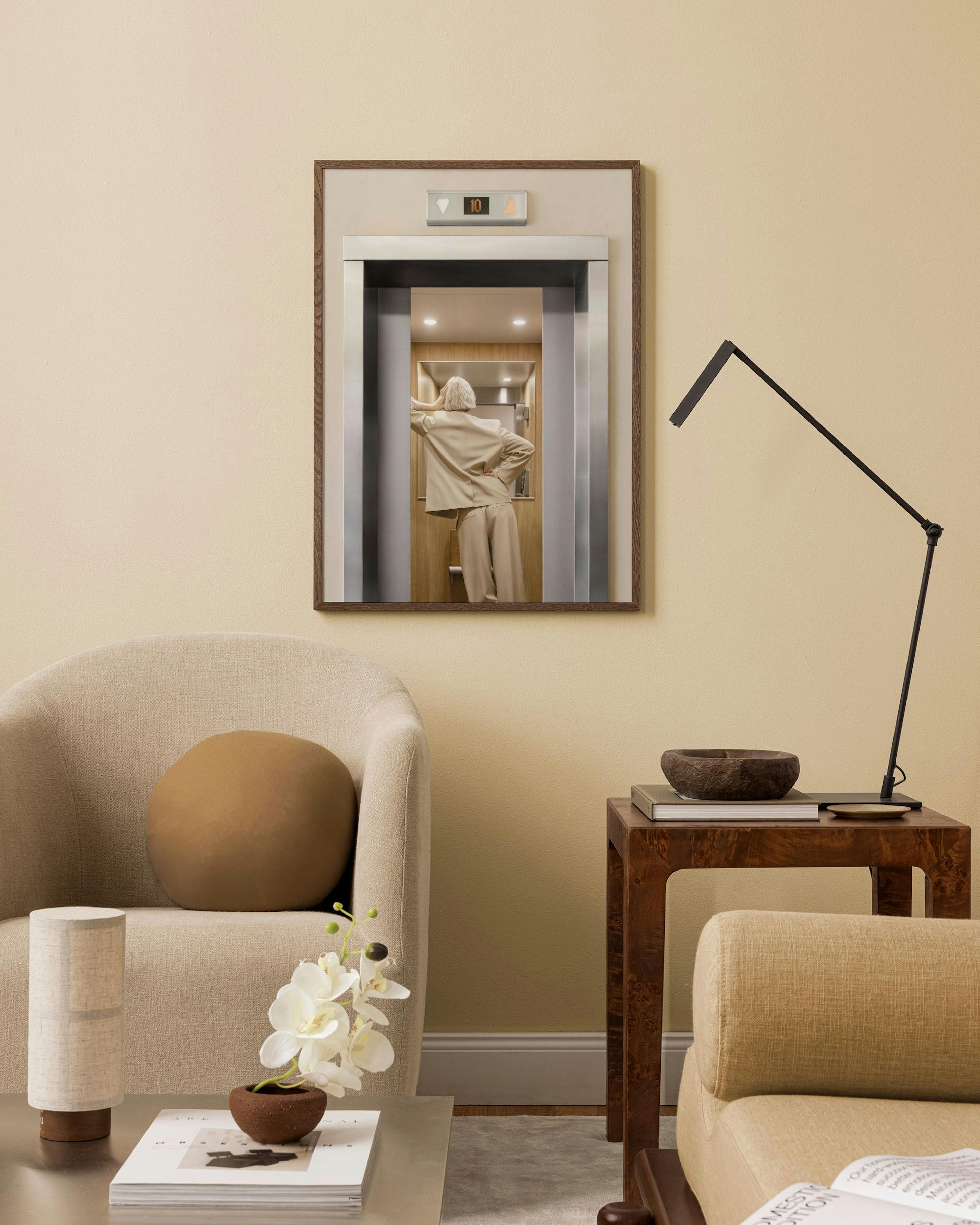 A print of a woman in an elevator, displayed on a beige wall above a beige armchair and a wooden side table.