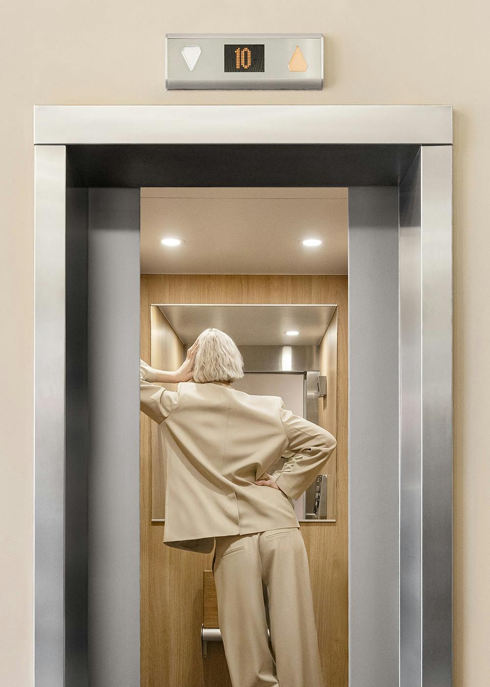 A print of a woman with blonde hair in a beige suit stepping into an open elevator with a digital display showing 10.