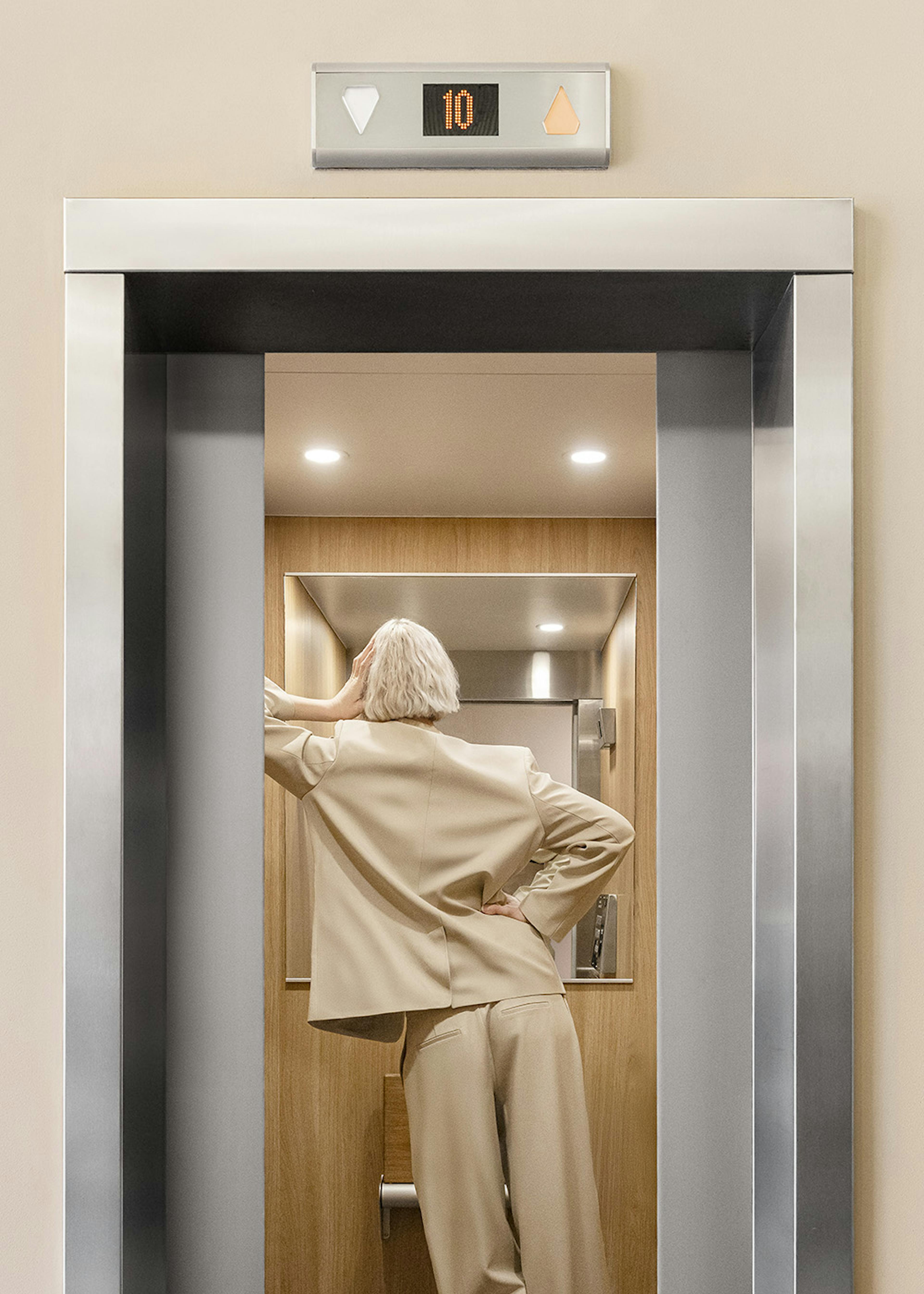 A print of a woman with blonde hair in a beige suit stepping into an open elevator with a digital display showing 10.