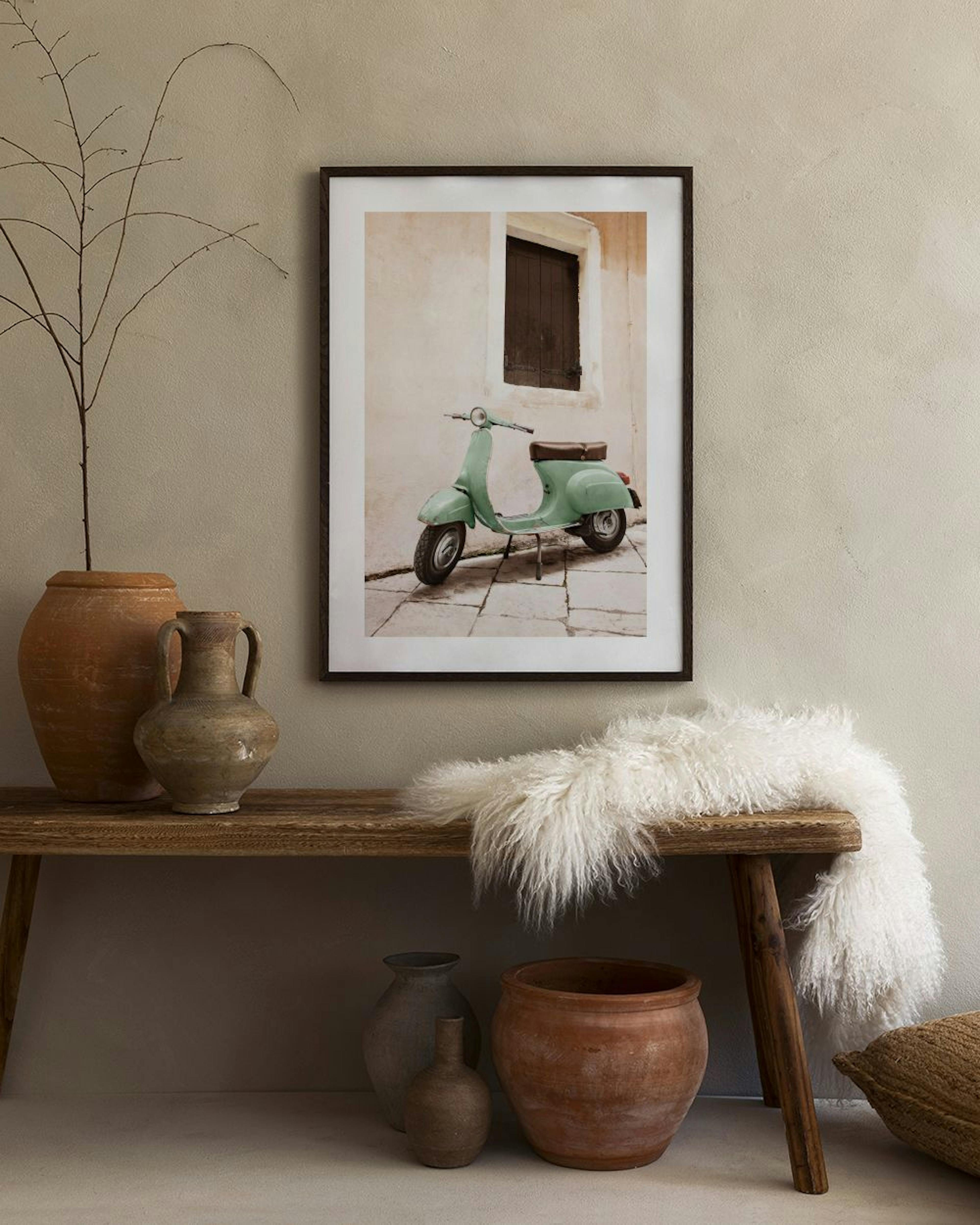 Poster di una vespa verde menta parcheggiata davanti a un muro color crema, appeso sopra un tavolino con vasi e libri.