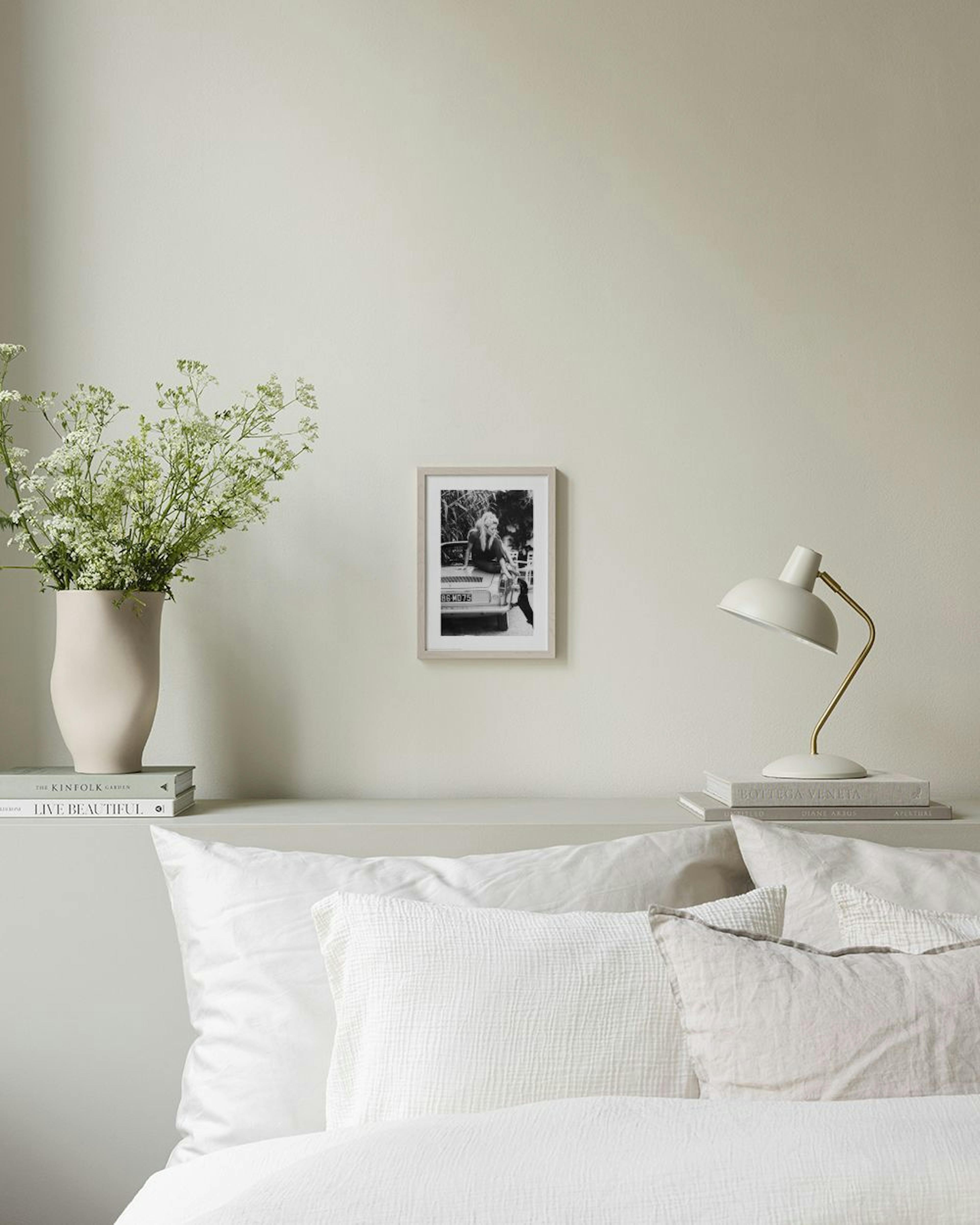 Schwarz-weiß Fotografie von Brigitte Bardot auf einem Auto, als Poster an einer grauen Wand, neben einem Stuhl und Vase.