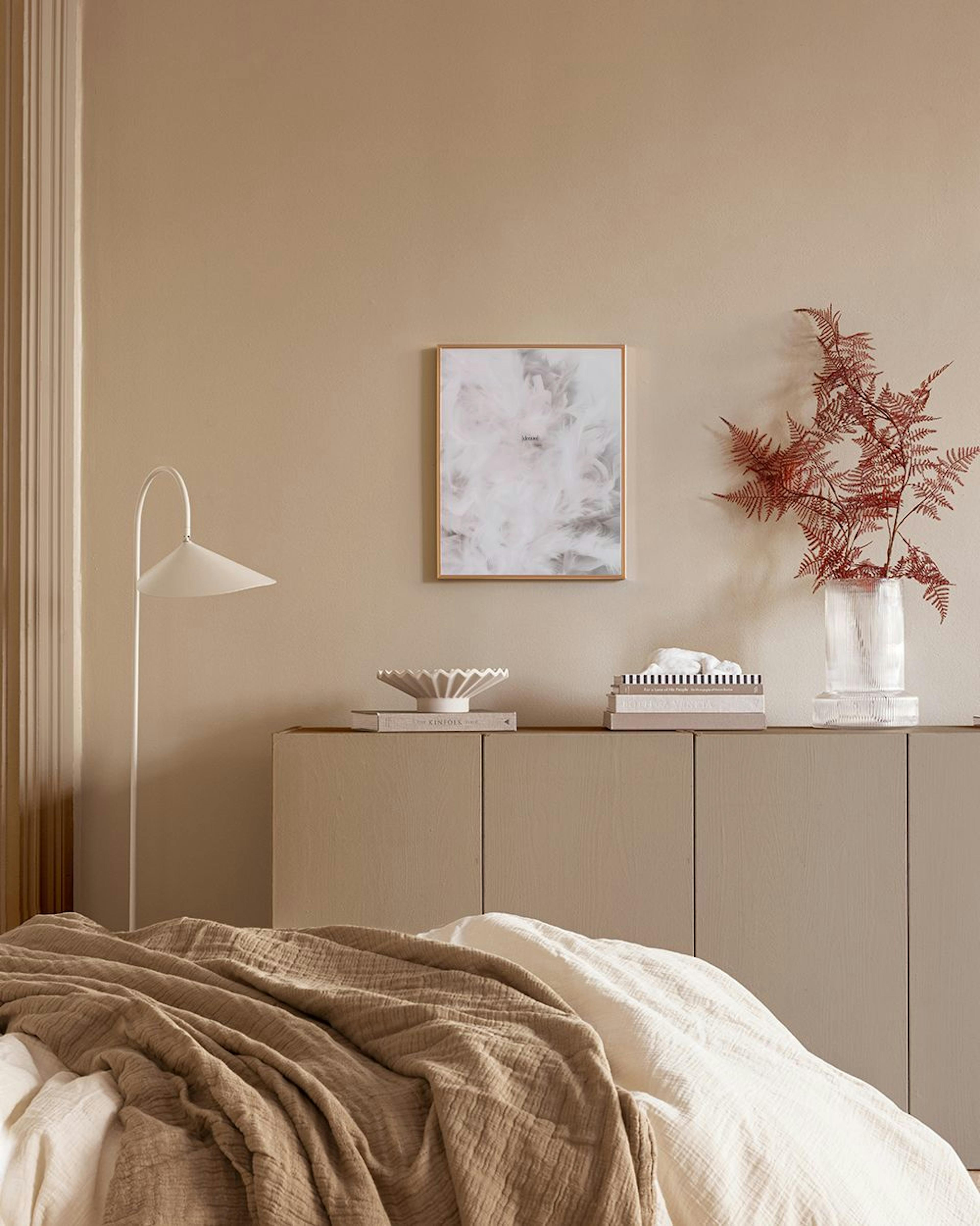 Póster de plumas blancas y grises, colgado en una cuadrícula negra sobre una pared blanca, junto a una silla y plantas.