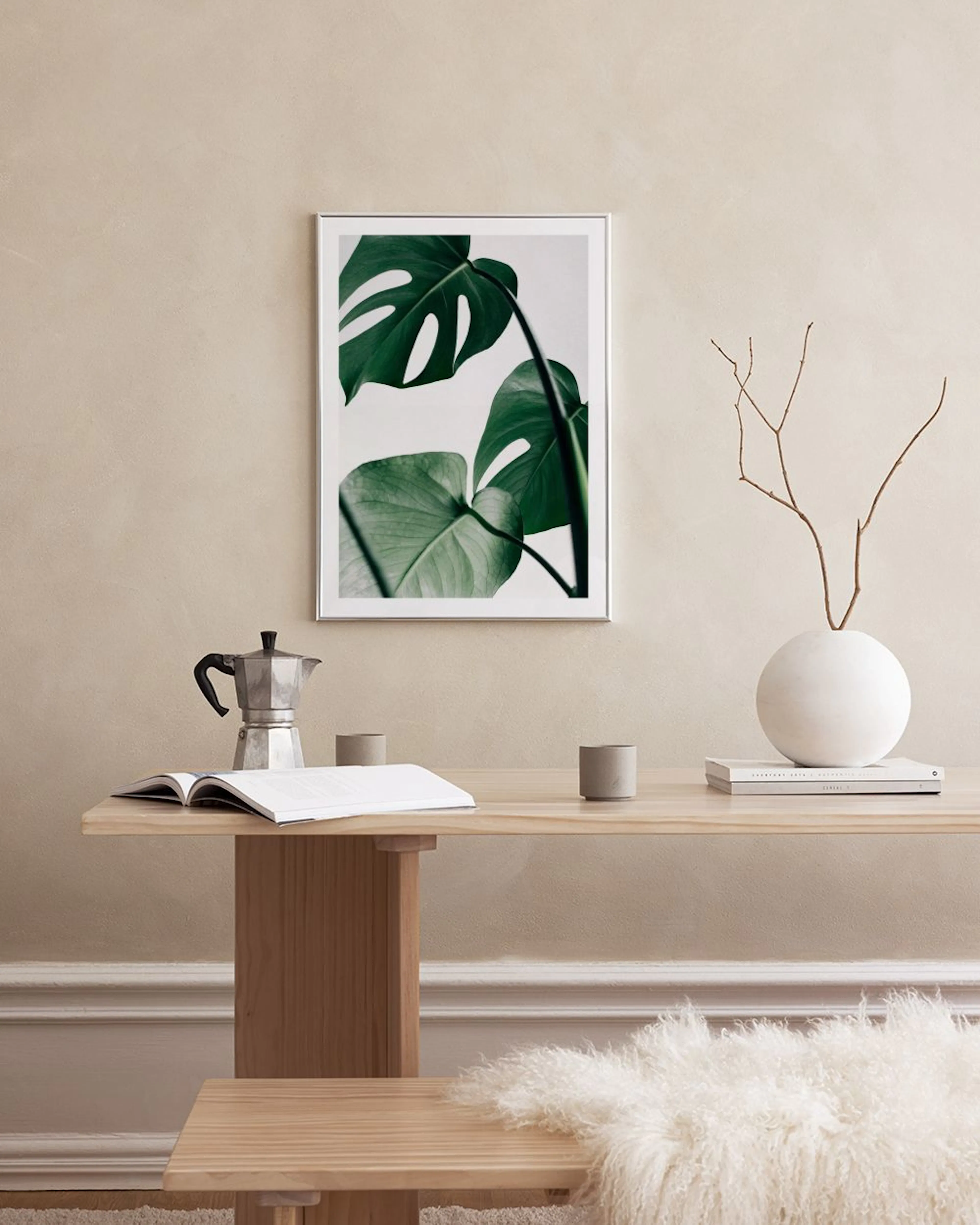 Plakat med grønne monstera blade på hvid baggrund, ophængt på et sort gitter i et minimalistisk rum med hvidt møblement.